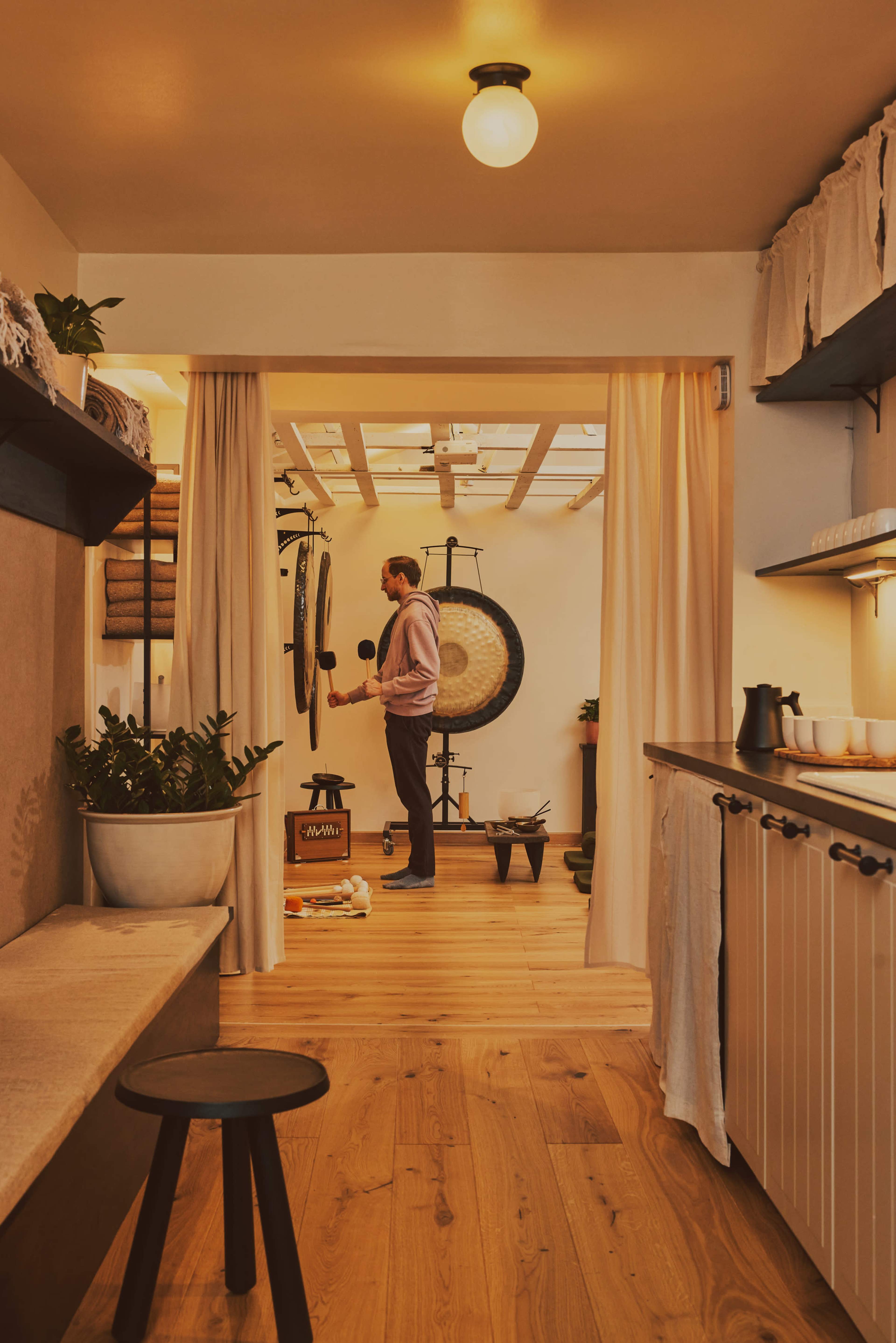 A person stands in a warmly lit room, playing a gong while surrounded by wooden decor and plants.