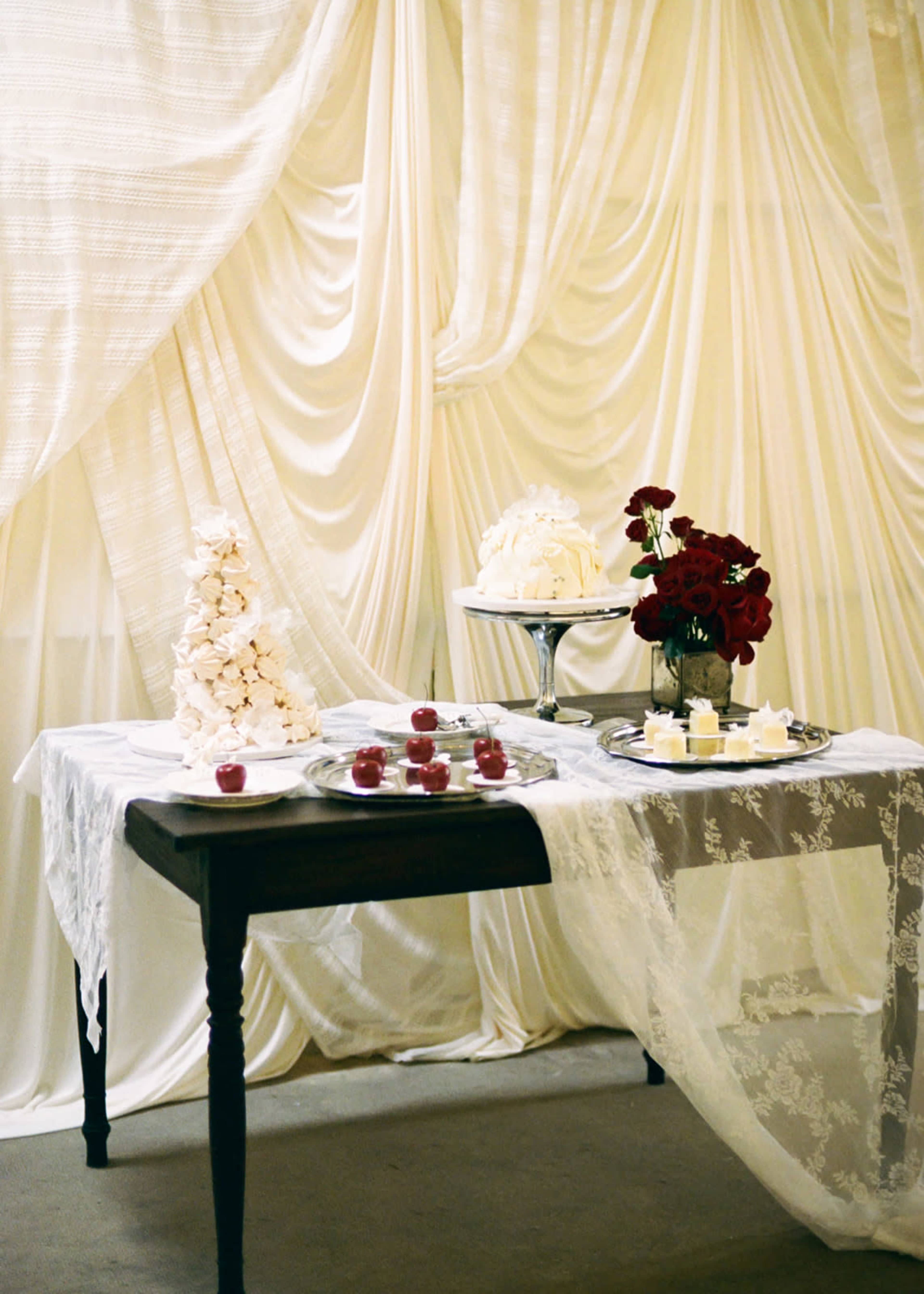 A table is adorned with a lace tablecloth and decorated with a floral arrangement, a cake, and small desserts against a backdrop of draped white fabric.