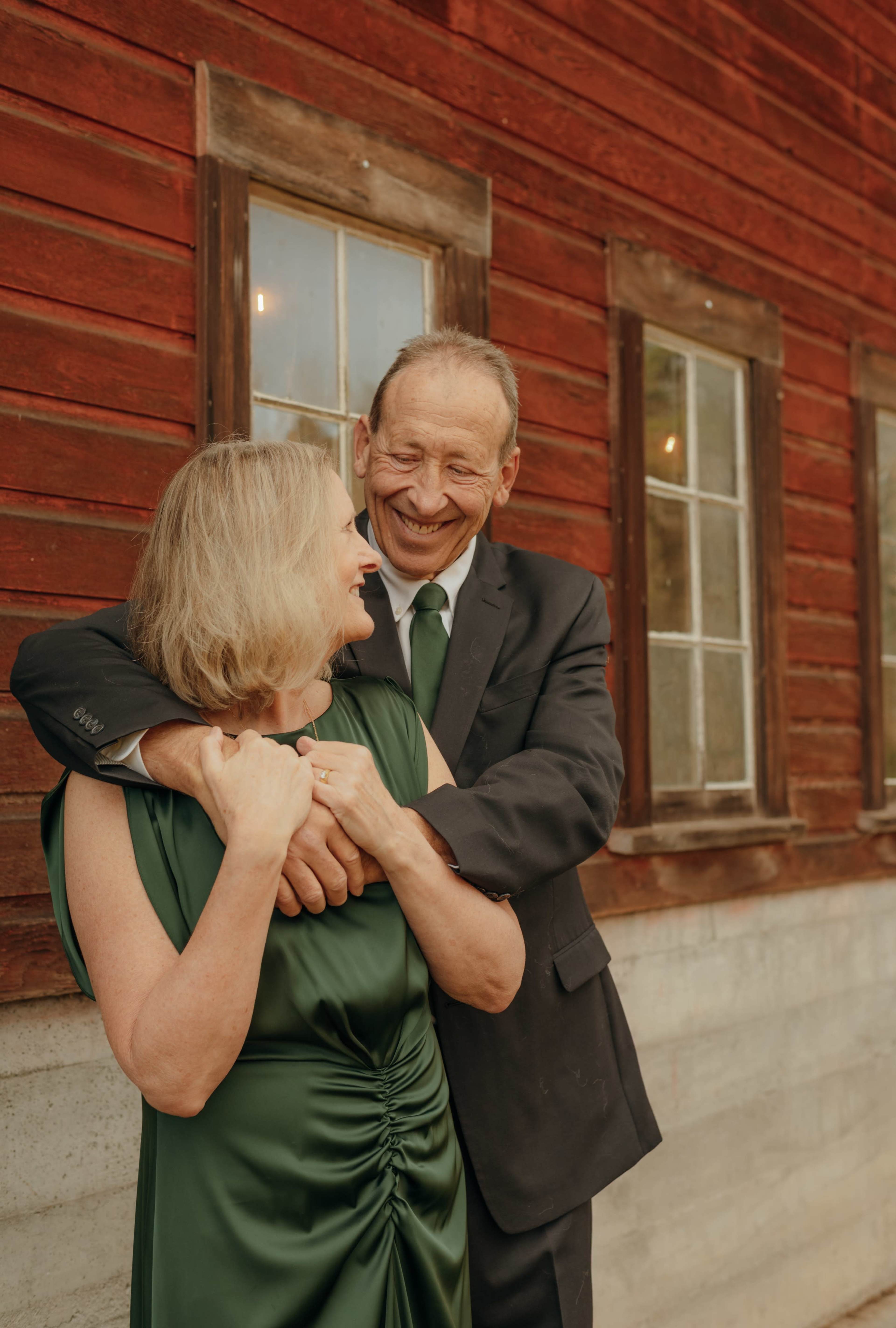 A man in a black suit embraces a woman in a green dress in front of a rustic red wooden building.