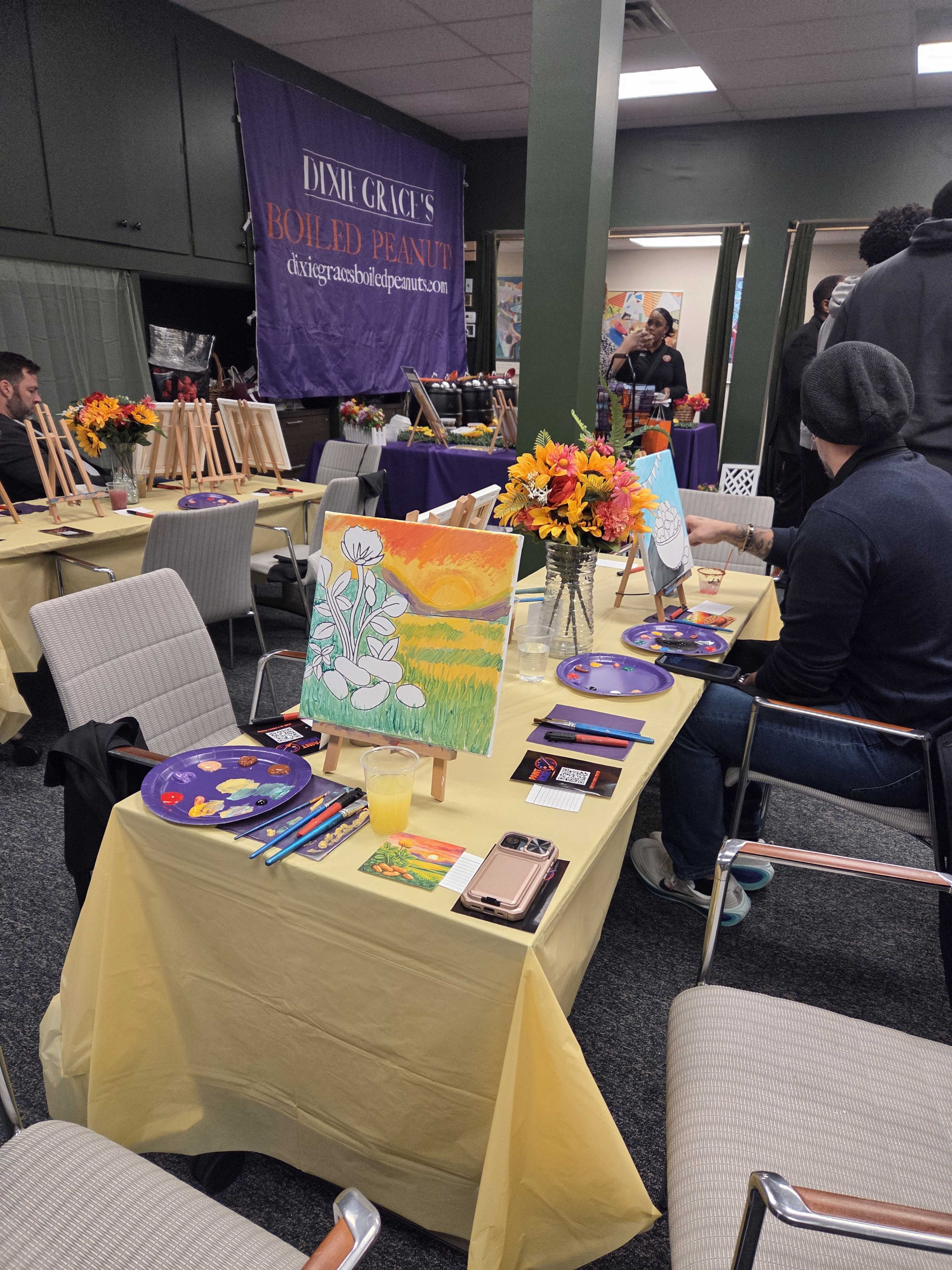 The image shows a bright indoor space set up for a painting event, featuring tables covered with yellow tablecloths, easels with canvases, and floral centerpieces.