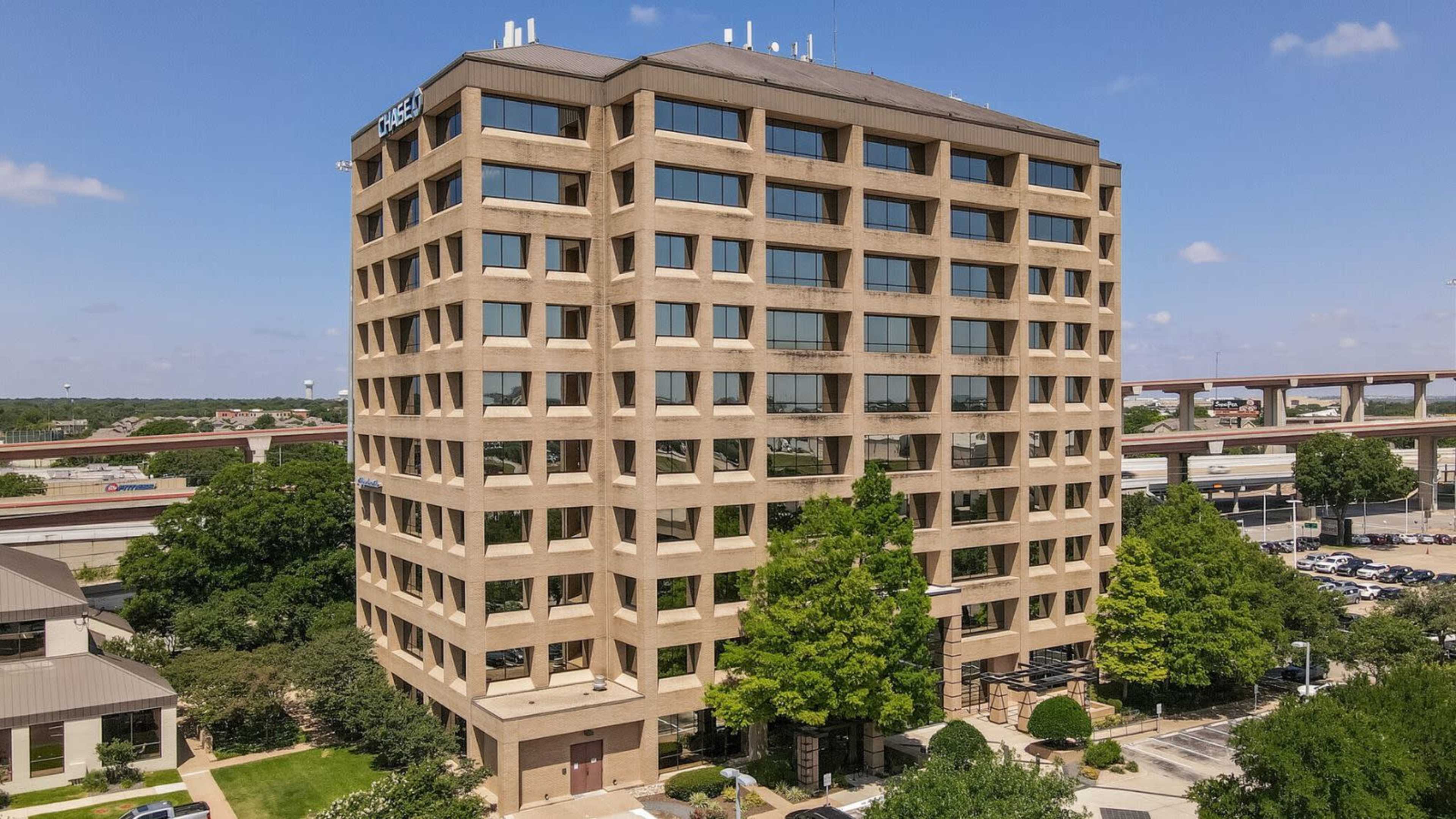 A multi-story commercial building with a flat roof and large windows, surrounded by greenery and parking spaces.