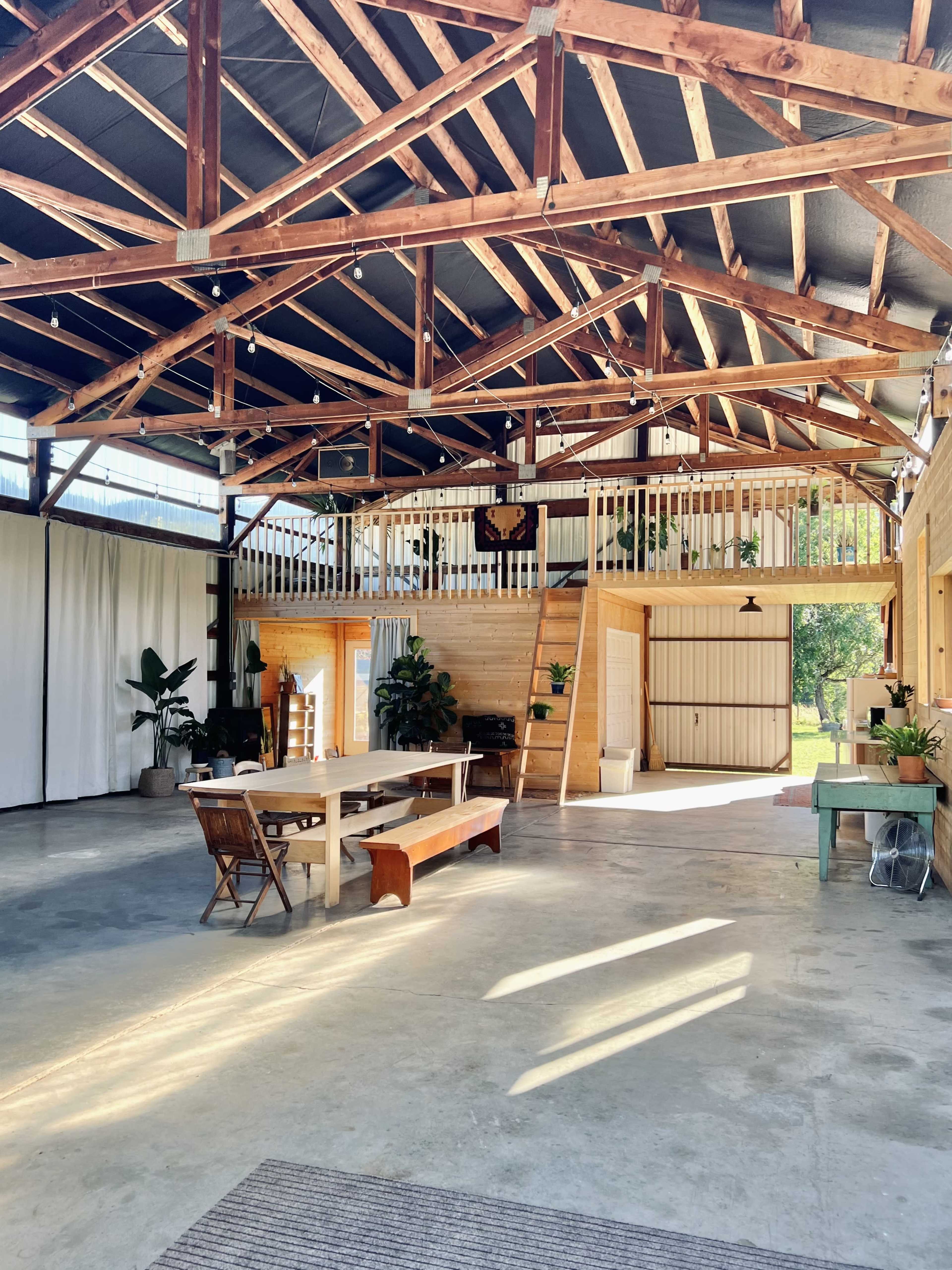 The image shows an open, spacious interior of a barn with a high wooden ceiling, featuring a long table and benches, along with various plants and light coming through large windows.