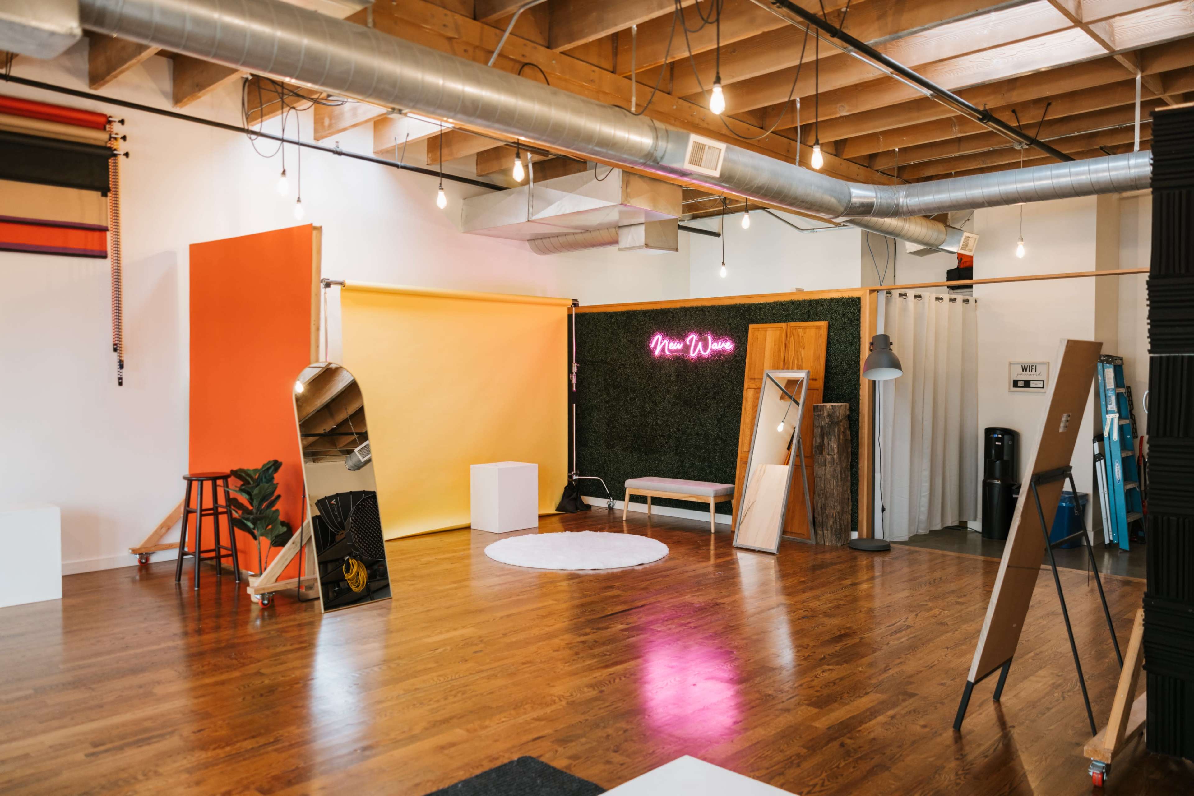 A spacious studio with wooden flooring, various backdrops including orange and yellow, a neon sign, and scattered furniture for photo shoots.
