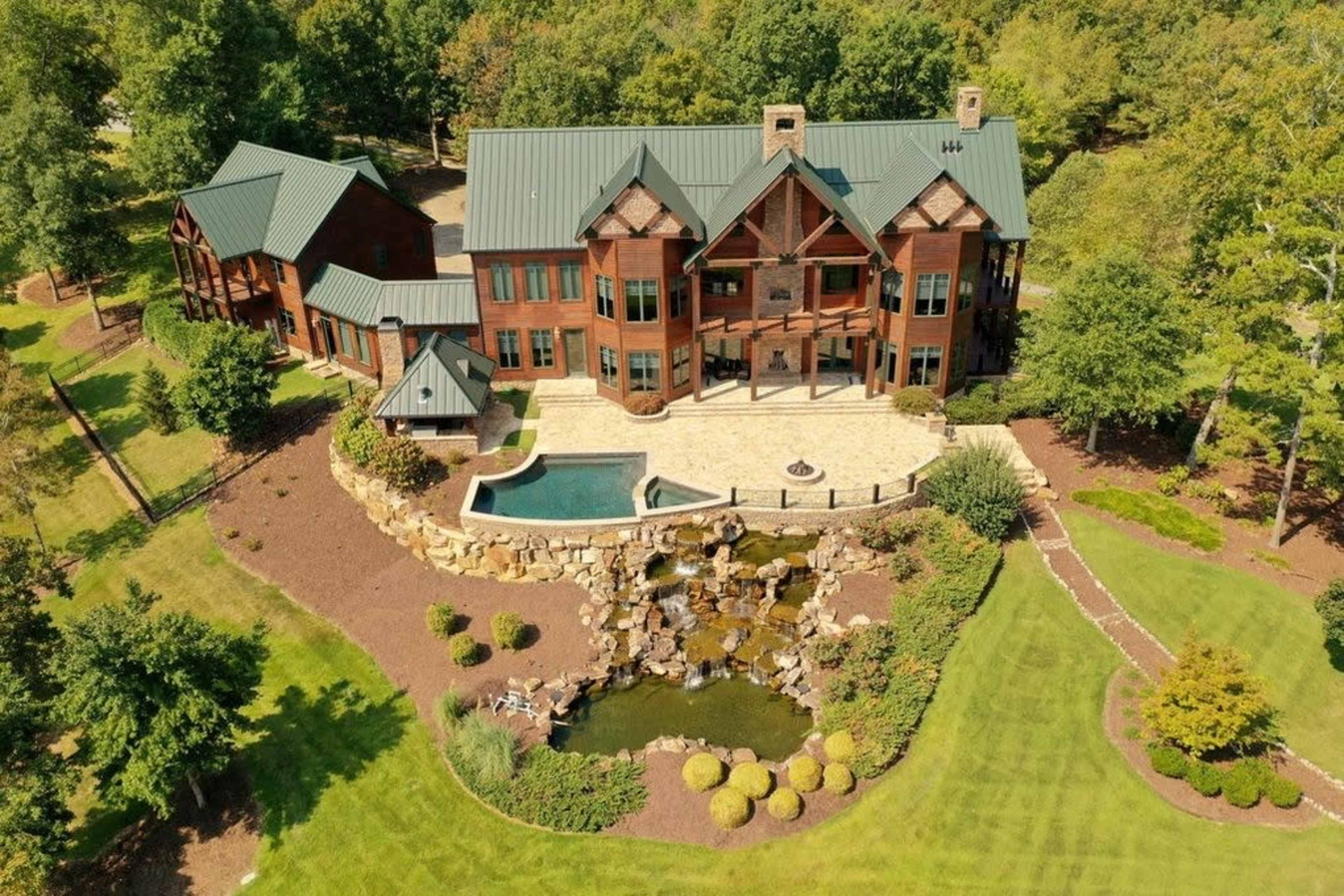 A large, multi-story wooden house with a green metal roof, surrounded by landscaped gardens and a pool with a cascading waterfall.