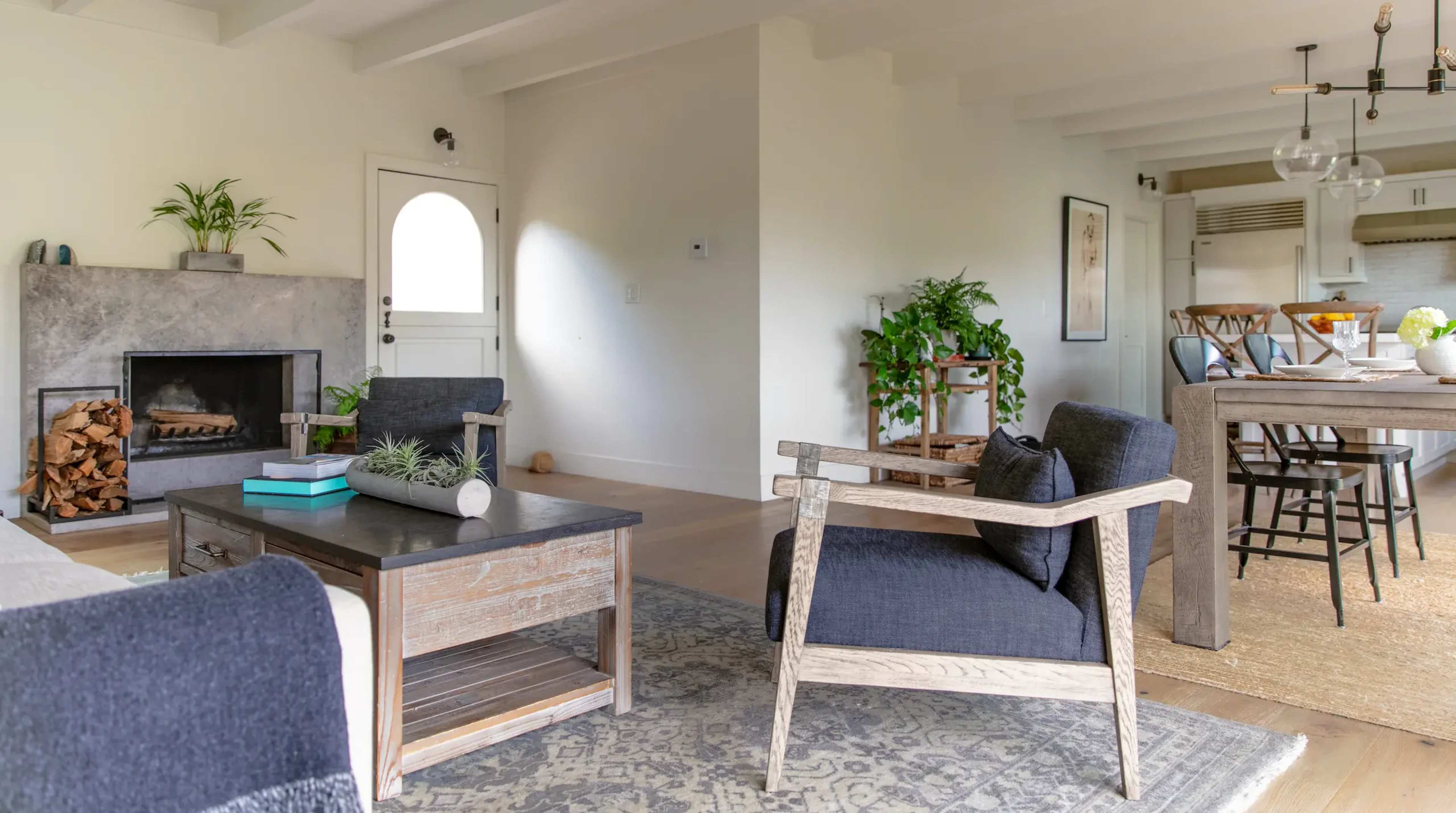 A modern living room featuring a fireplace, two armchairs, a coffee table, and plants, with an open view into a kitchen area.