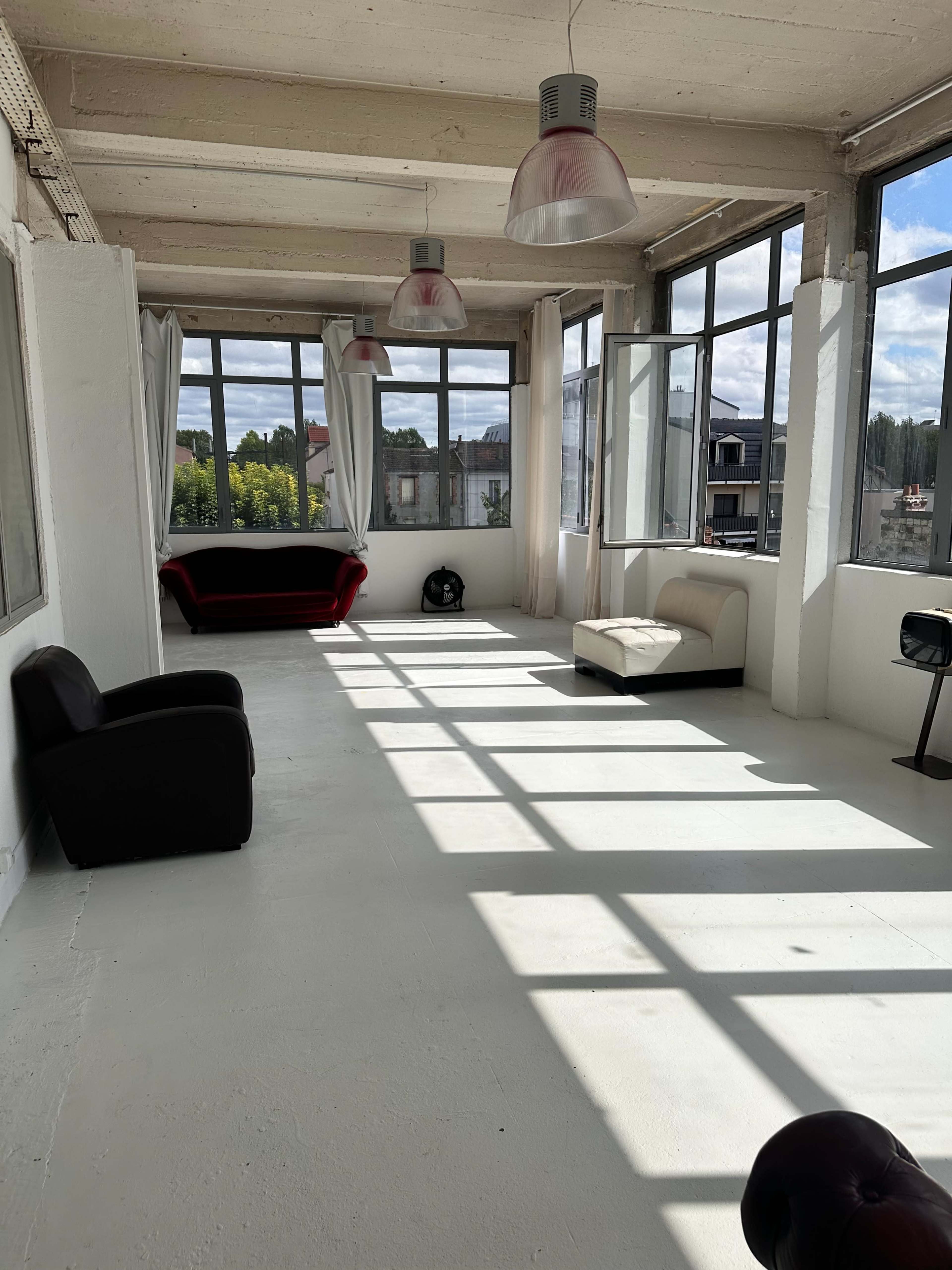 The image shows a spacious room with large windows, concrete walls, and a few pieces of furniture, including a red sofa, a white couch, and a black chair.