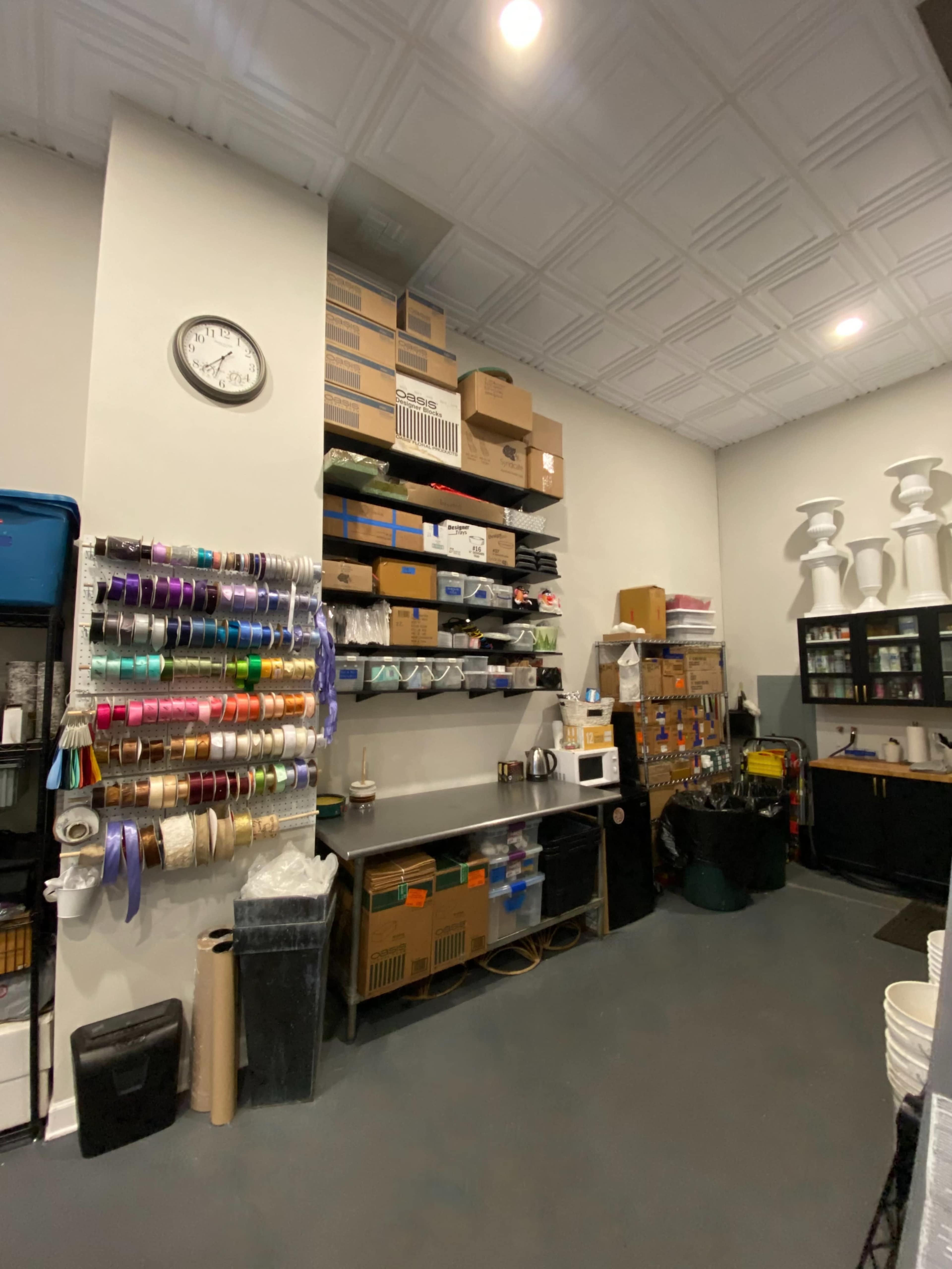 A storage area with organized shelves containing boxes, ribbons, and various supplies, alongside a clock on the wall and a small kitchenette.