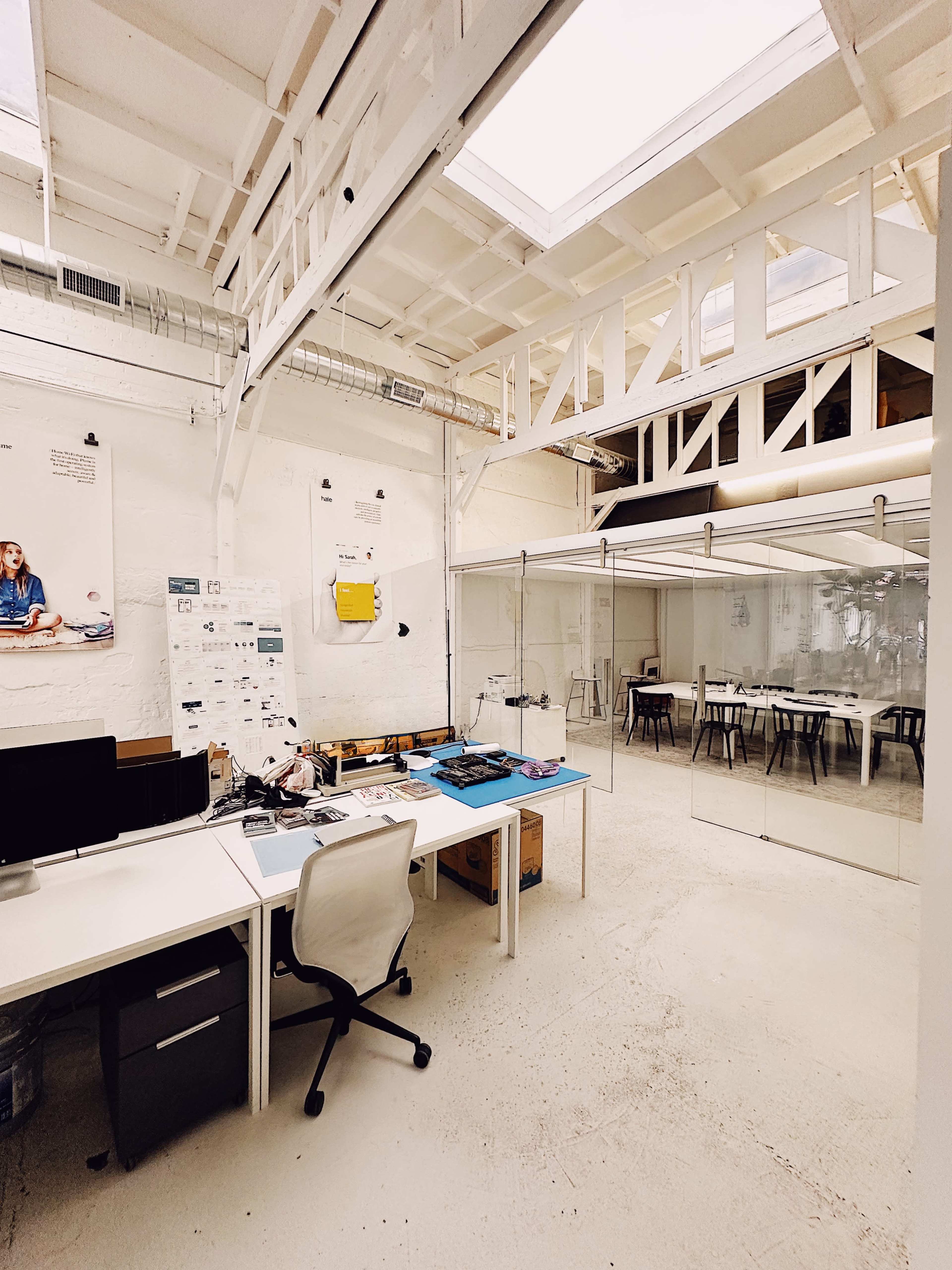 A spacious office with a white interior, featuring a desk area with a chair, glass-walled meeting rooms, and tables in a separate section.