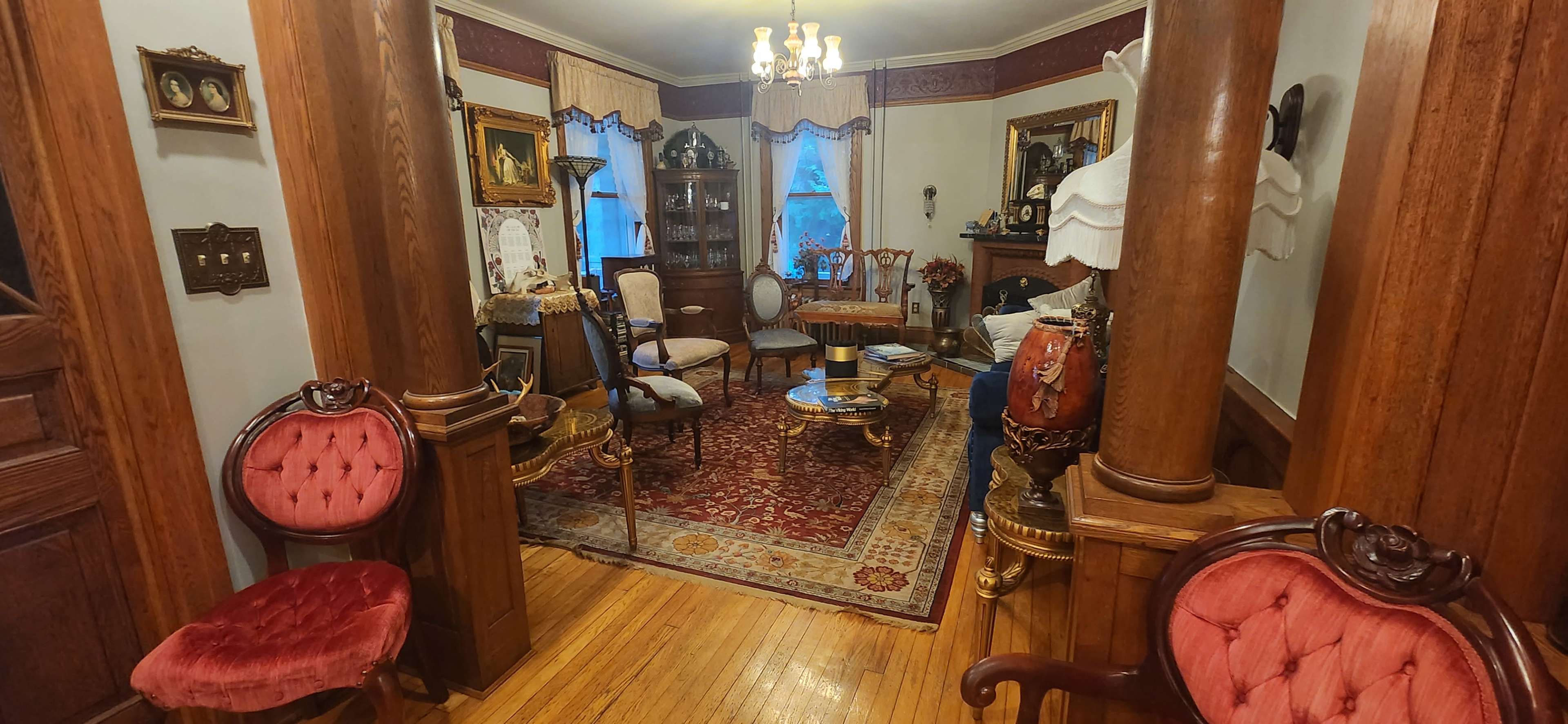 The image shows a vintage living room with wooden pillars, a patterned area rug, ornate furniture, and framed artwork on the walls.