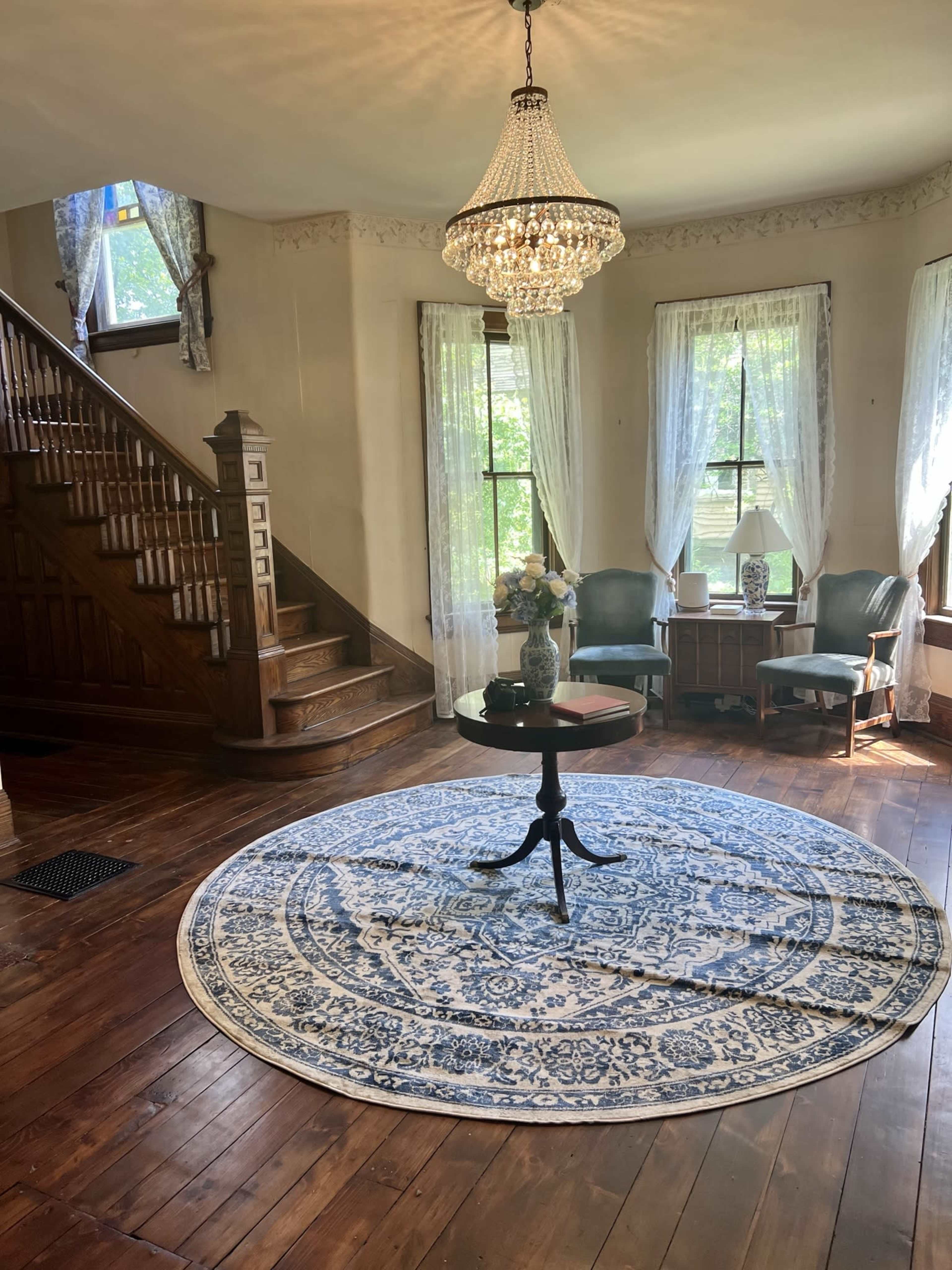 A spacious living area with a circular rug, a chandelier overhead, and a staircase in the background, illuminated by natural light through large windows.