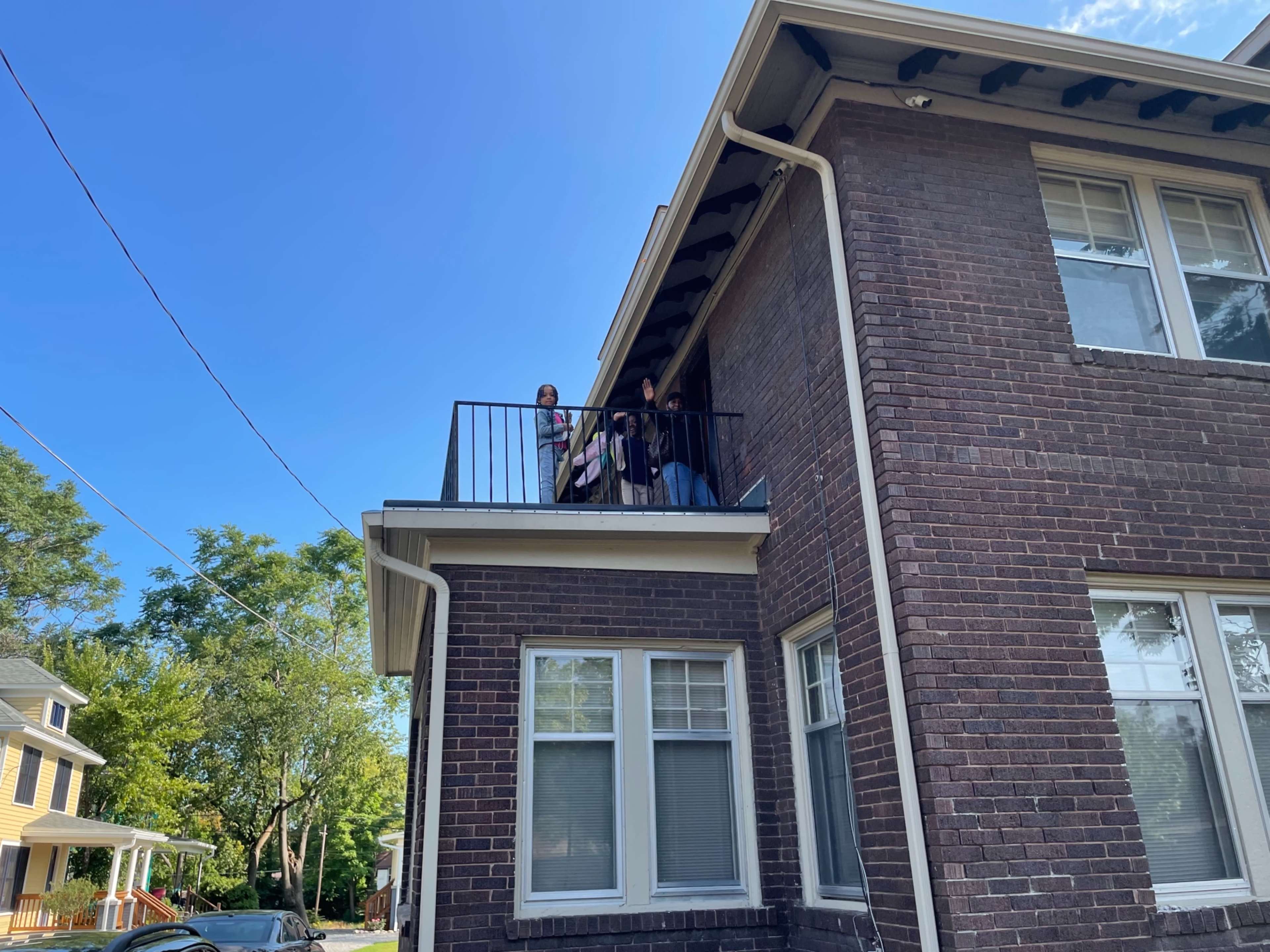 Two people stand on a balcony of a brick apartment building on a sunny day.