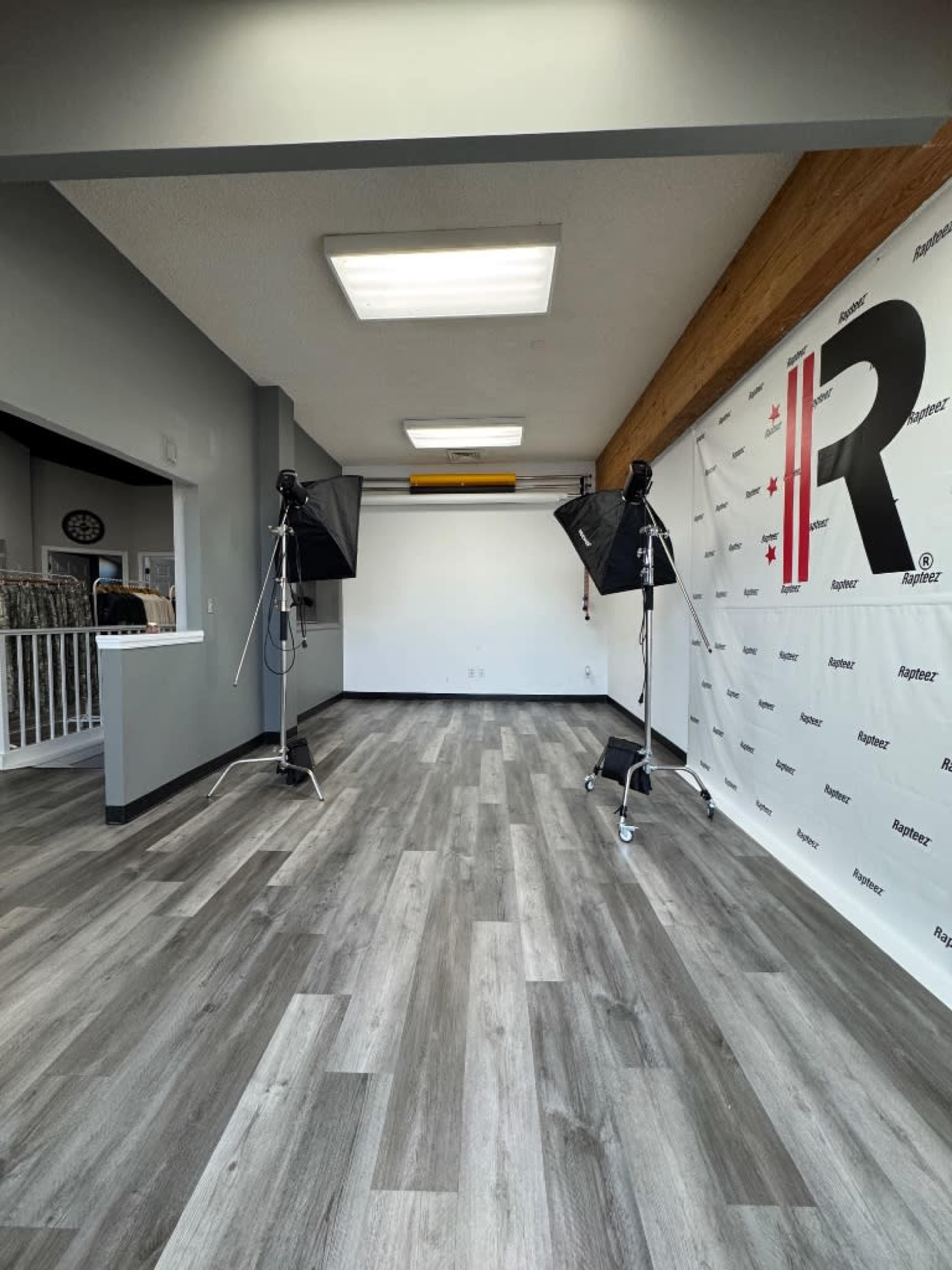The image shows a well-lit interior space featuring a plain backdrop with a logo, two studio lights on stands, and gray laminate flooring.