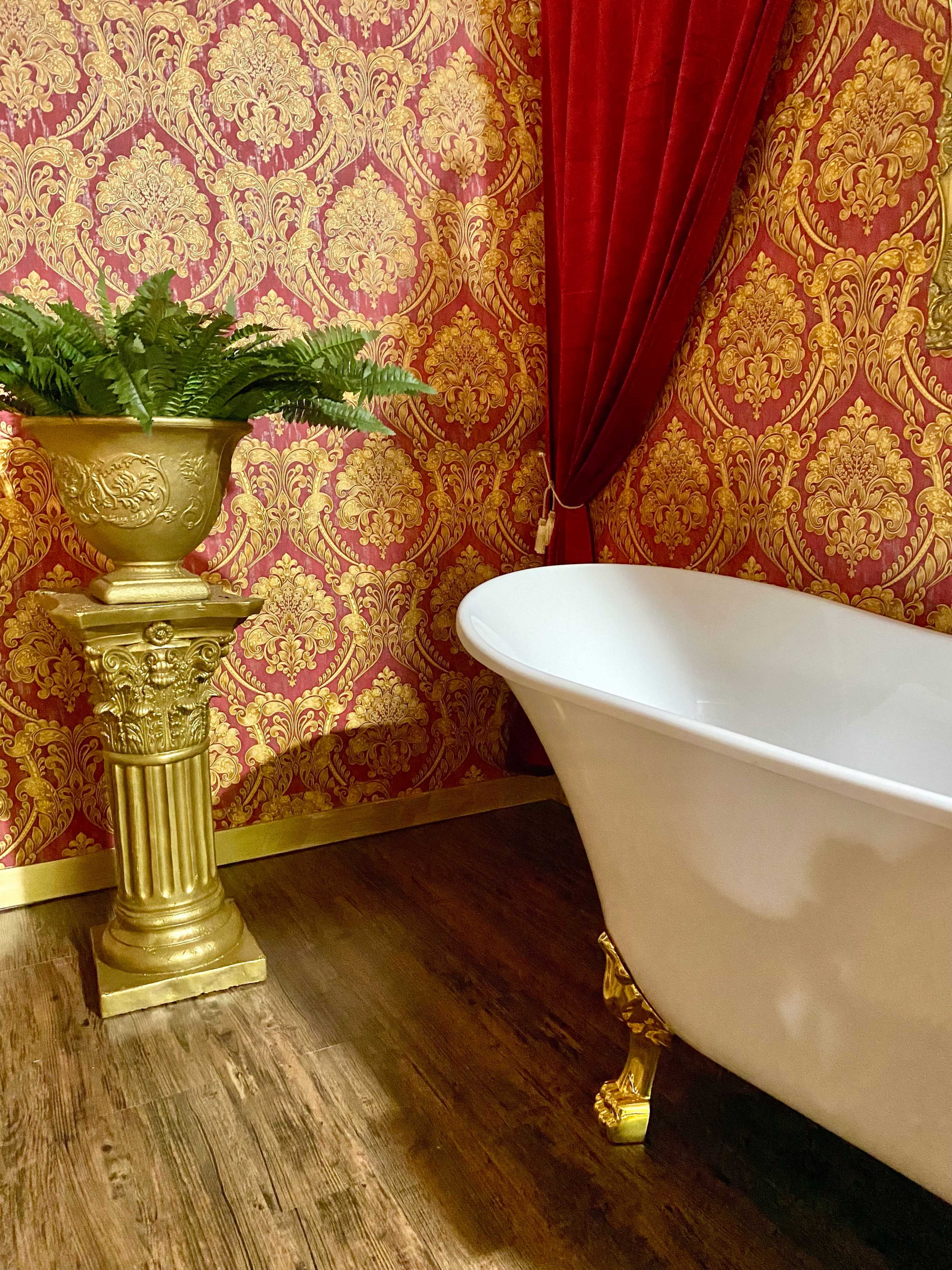 A white clawfoot bathtub is positioned next to a decorative gold pedestal holding a fern, against a backdrop of ornate red and gold wallpaper.