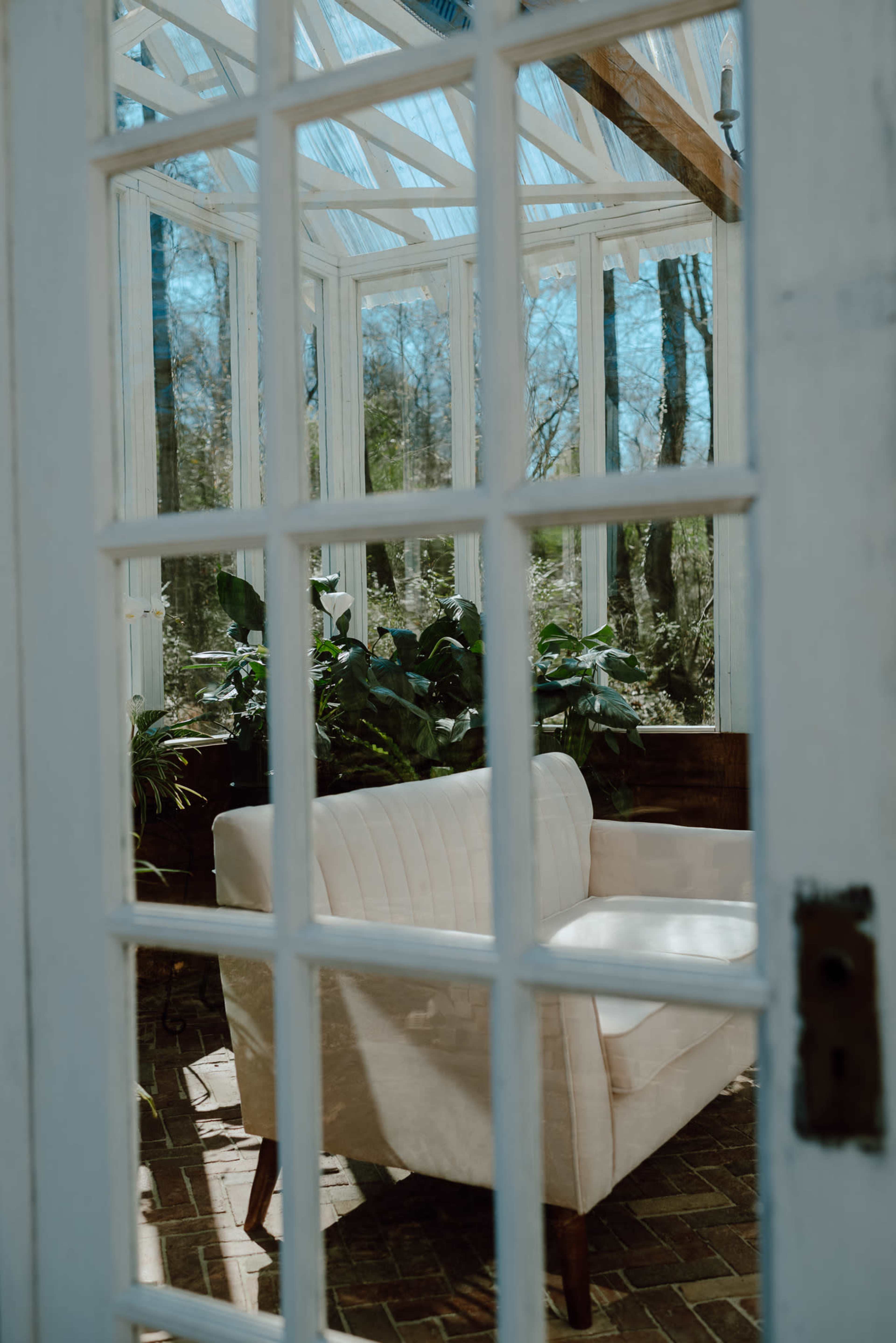 A light-colored armchair is visible through a glass door, set inside a bright greenhouse surrounded by potted plants.