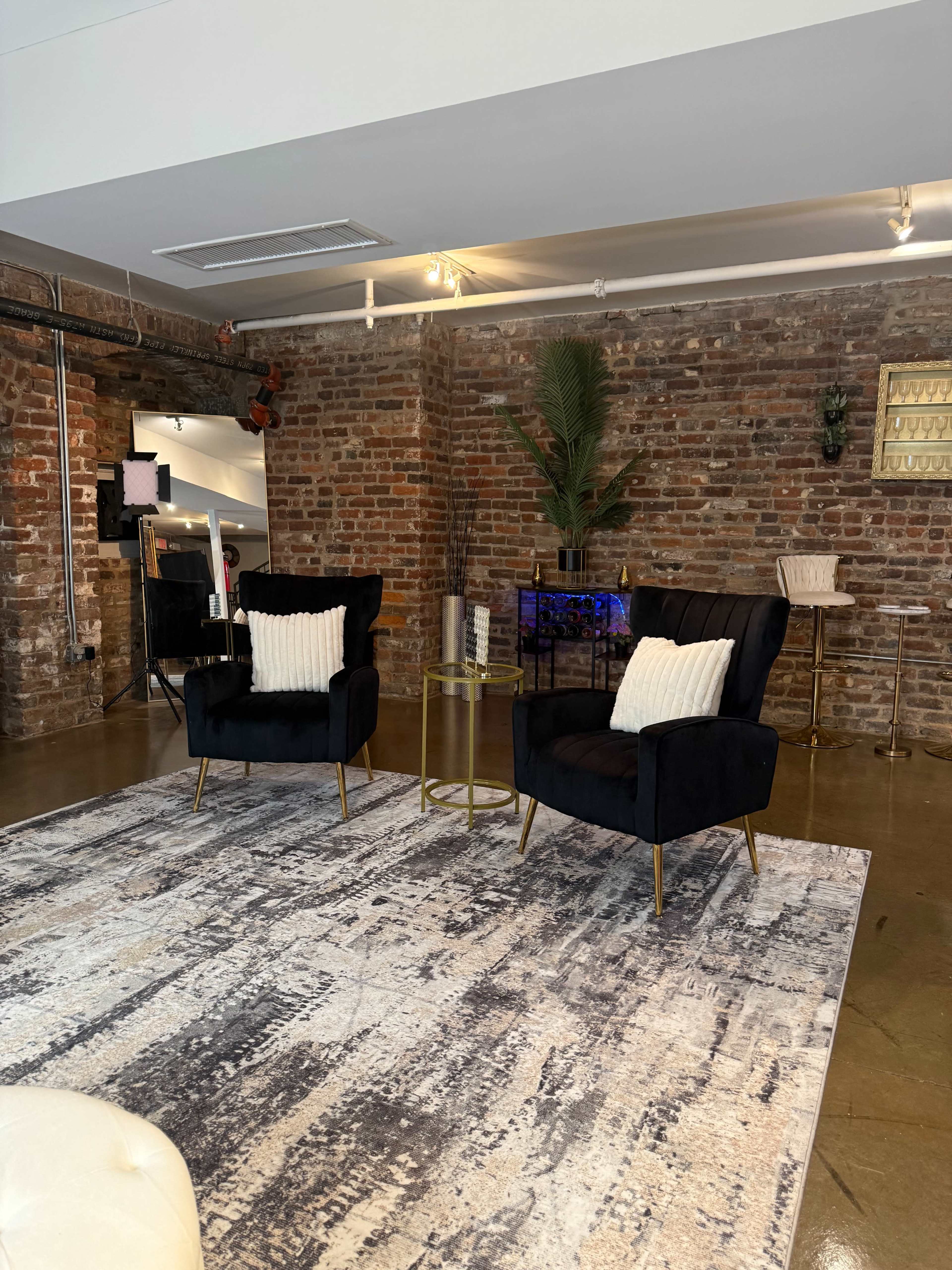 The image shows a modern interior with two black chairs and a small gold table on a patterned rug, set against a backdrop of exposed brick walls.