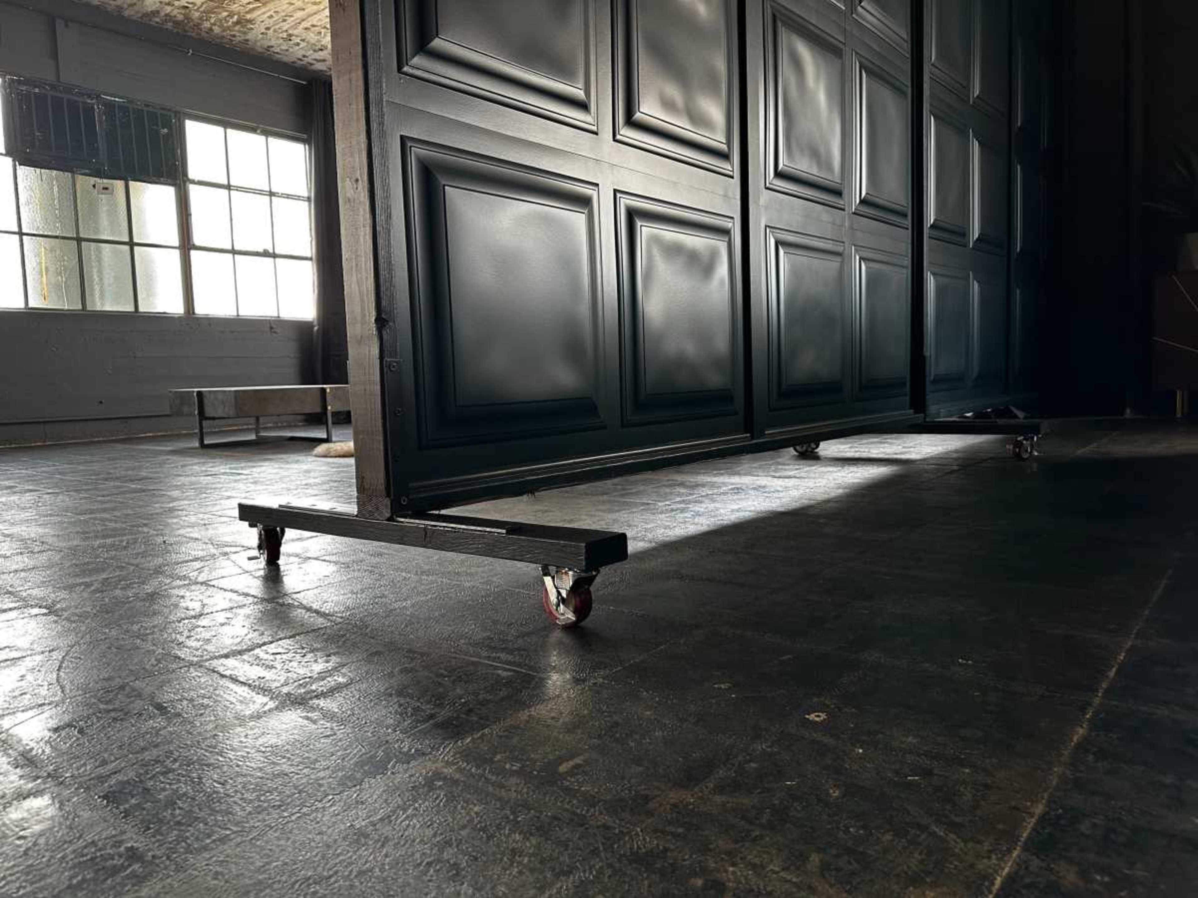 A large, closed black garage door is positioned on a wheeled track in an industrial-style interior with a window in the background.
