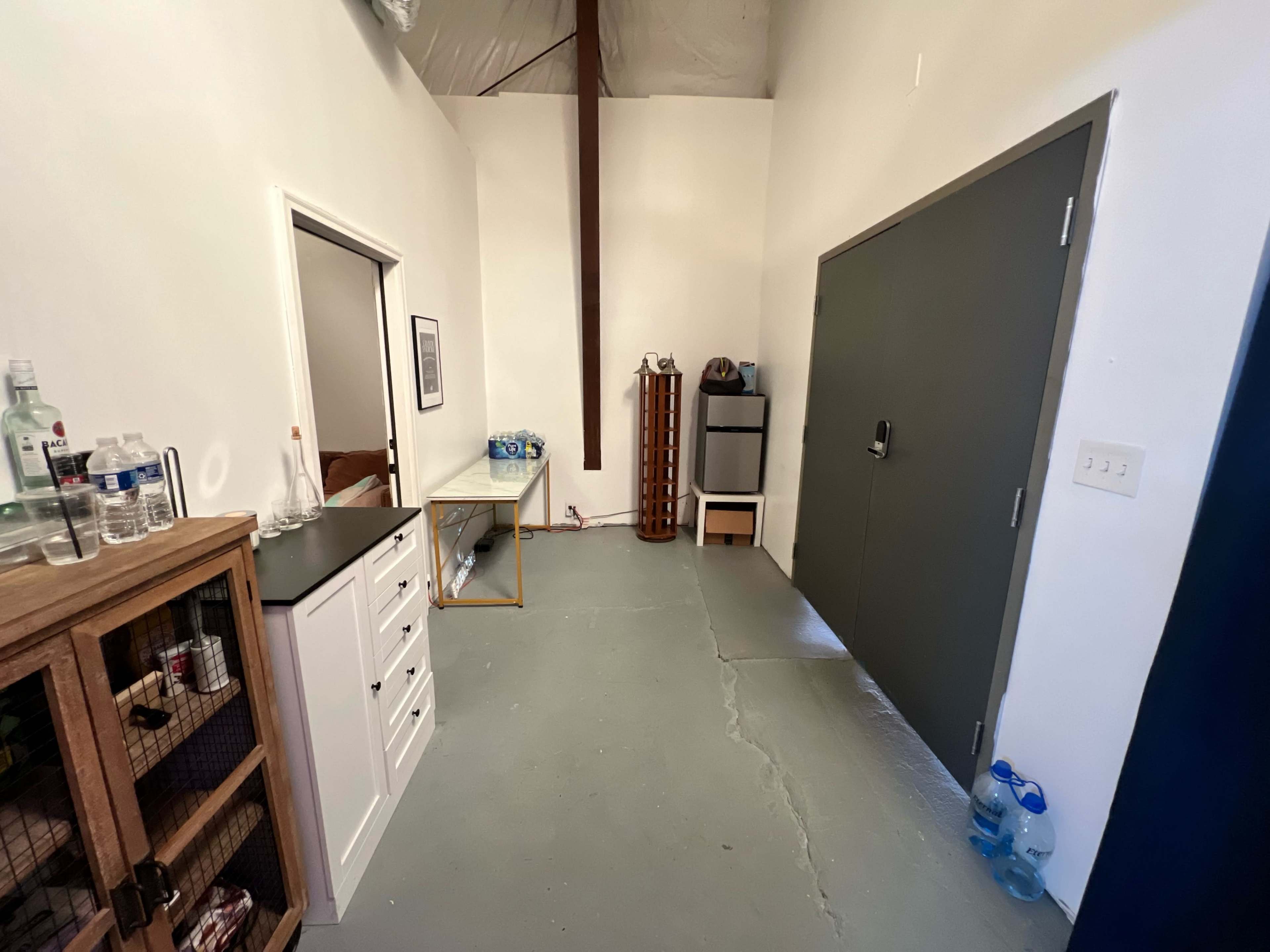 The image shows a spacious room with gray concrete flooring, featuring a large door on one side, a dresser, and a table with various items on it against the wall.
