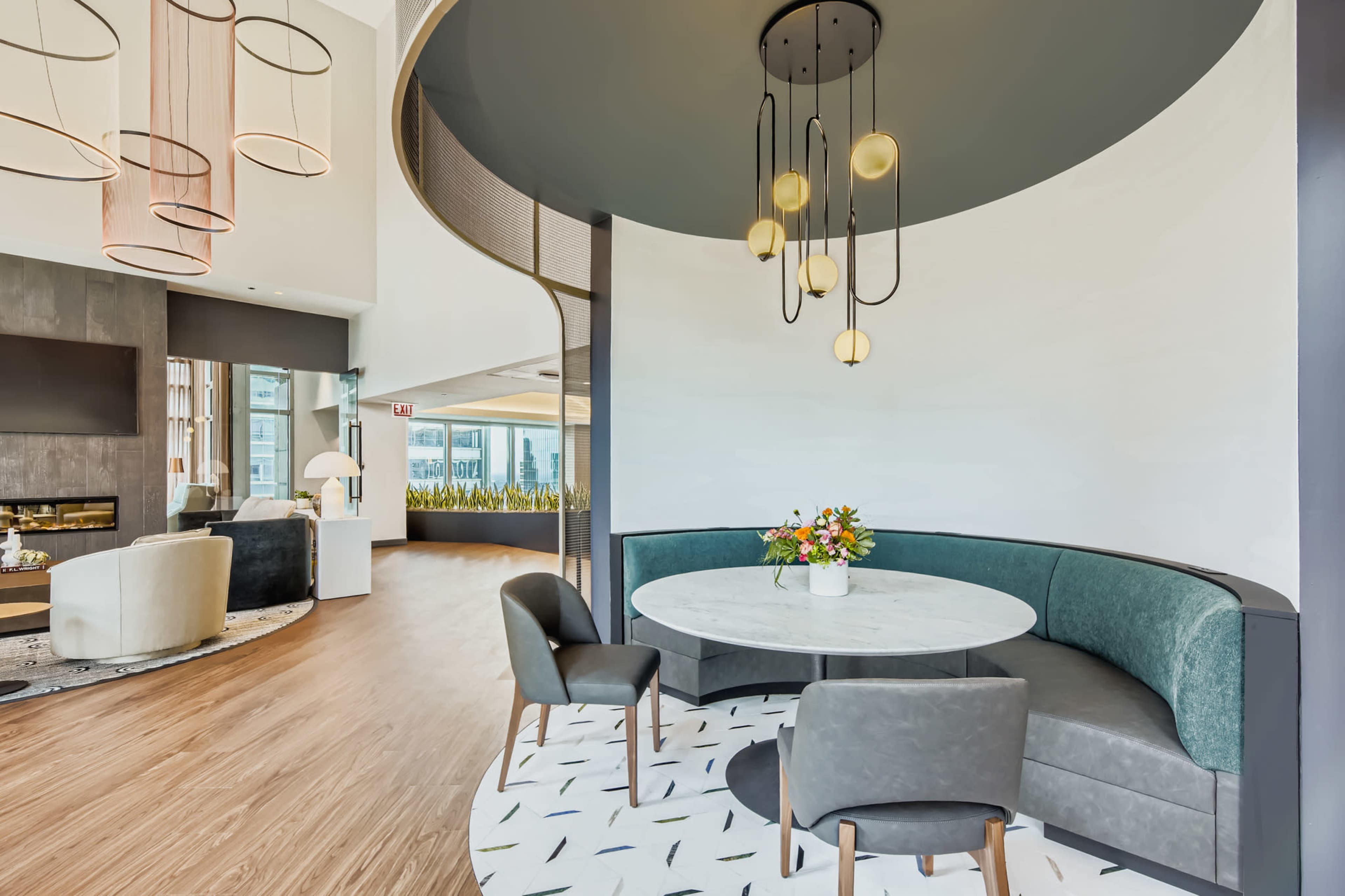 The image features a modern dining area with a circular marble table, upholstered seating, and a decorative light fixture, surrounded by neutral-toned walls and wooden flooring.