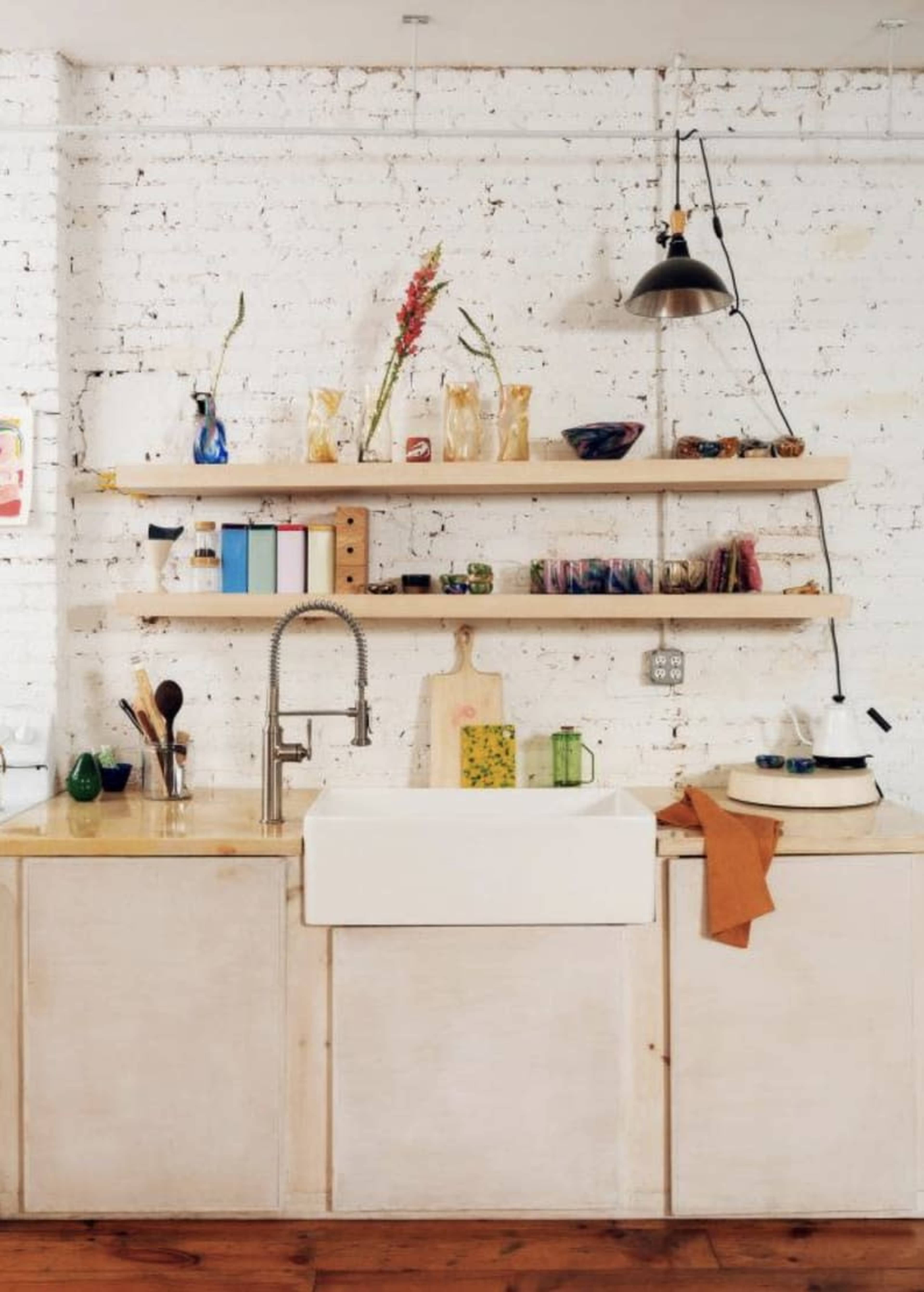 A kitchen features a large farmhouse sink with open shelving above displaying various kitchenware and decorative items against a white brick wall.
