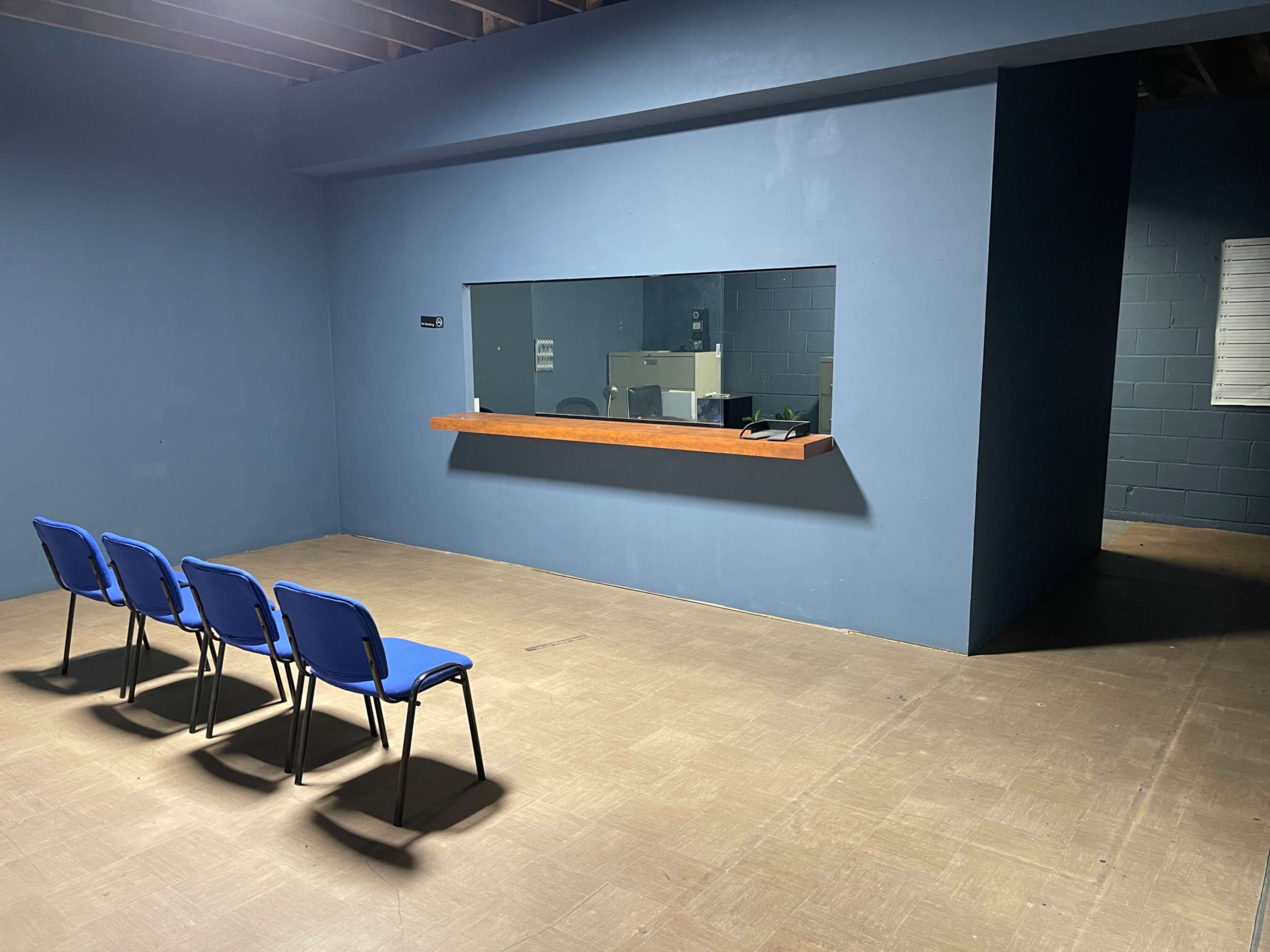 An empty room features four blue chairs arranged in a row facing a large window embedded in a blue wall.
