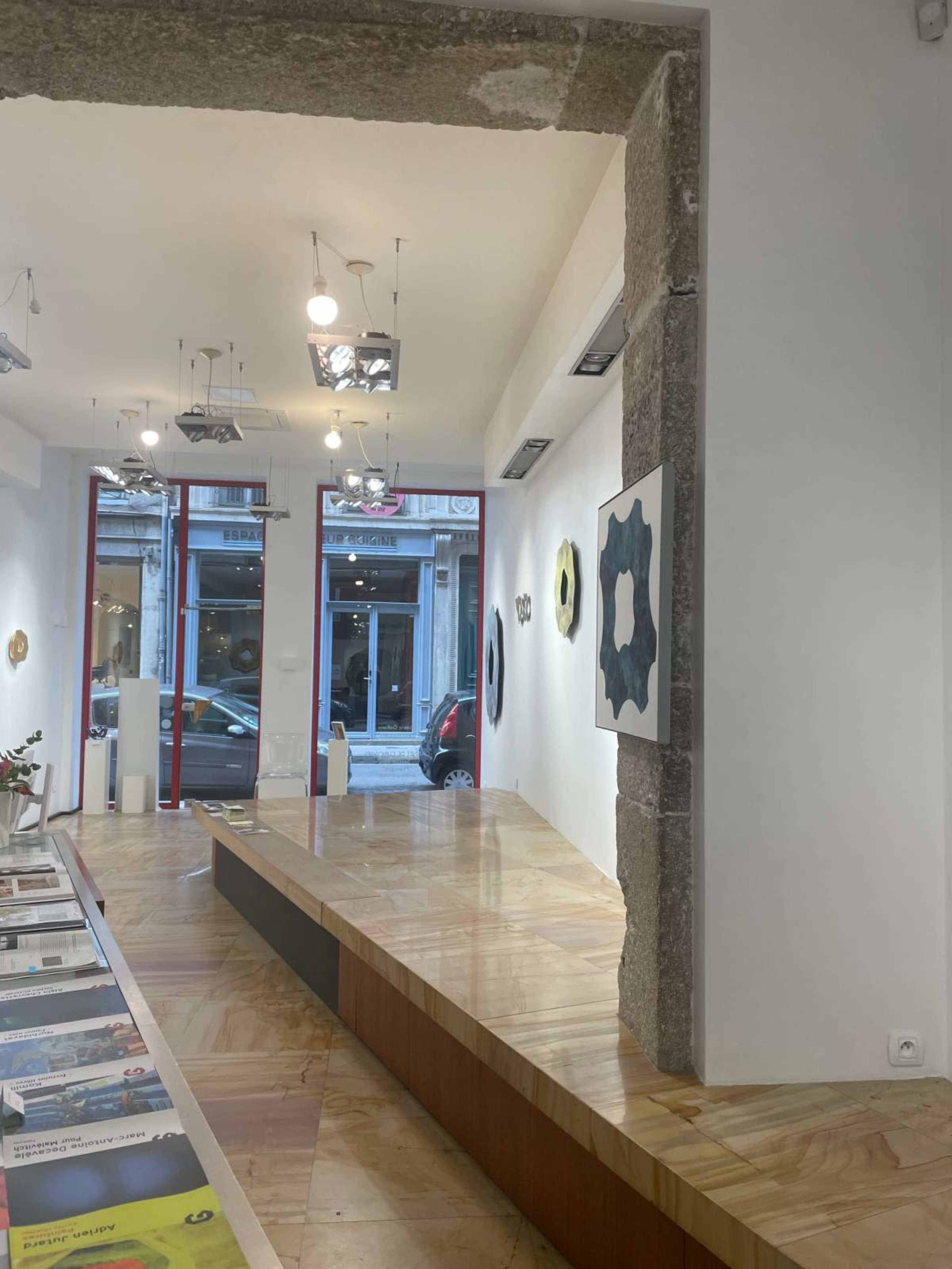 The image shows an interior of an art gallery with a wooden floor, illuminated by hanging lights, and featuring artworks on the walls alongside a reception area with brochures.
