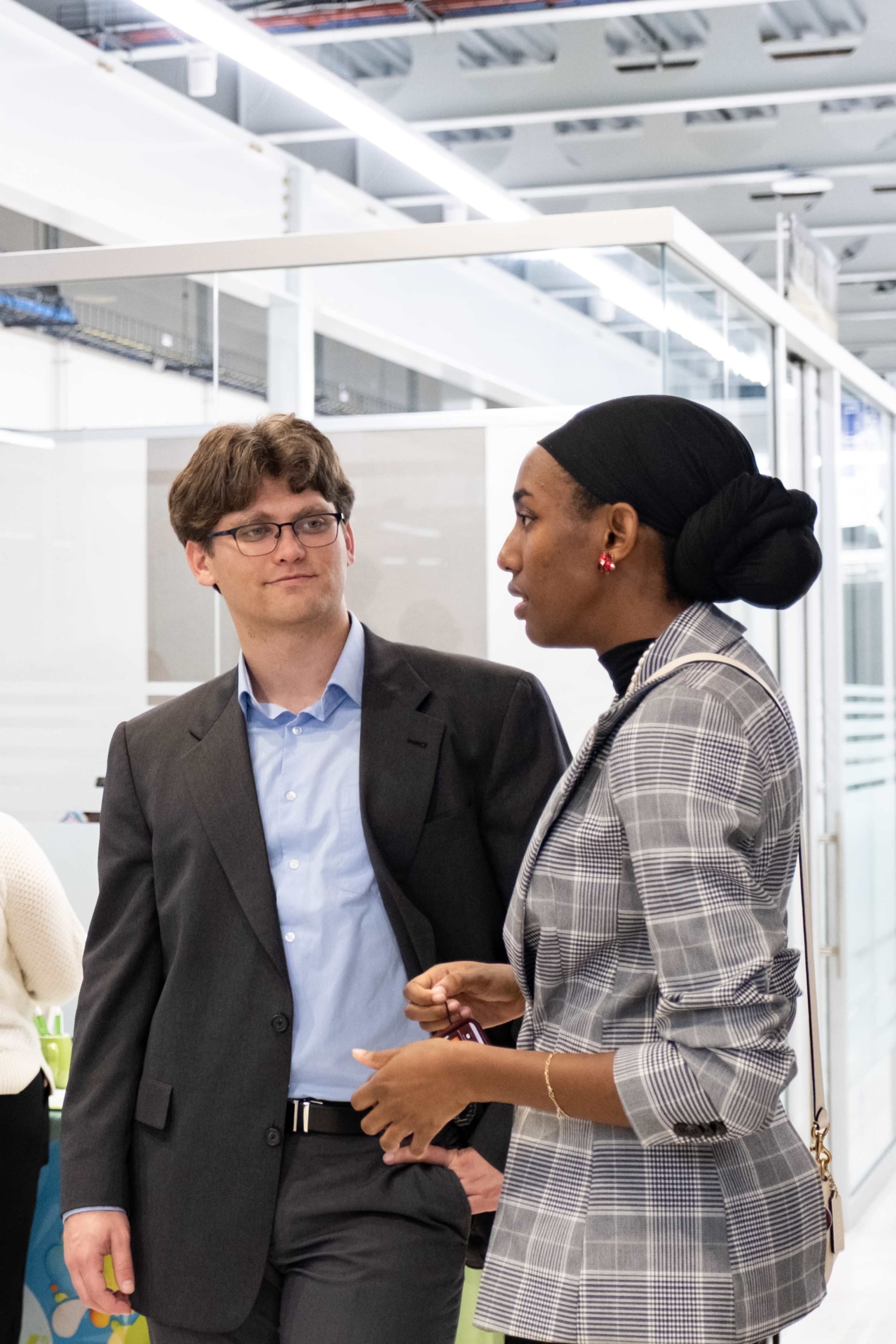 A man in a suit engages in conversation with a woman dressed in a checkered blazer in a modern office environment.