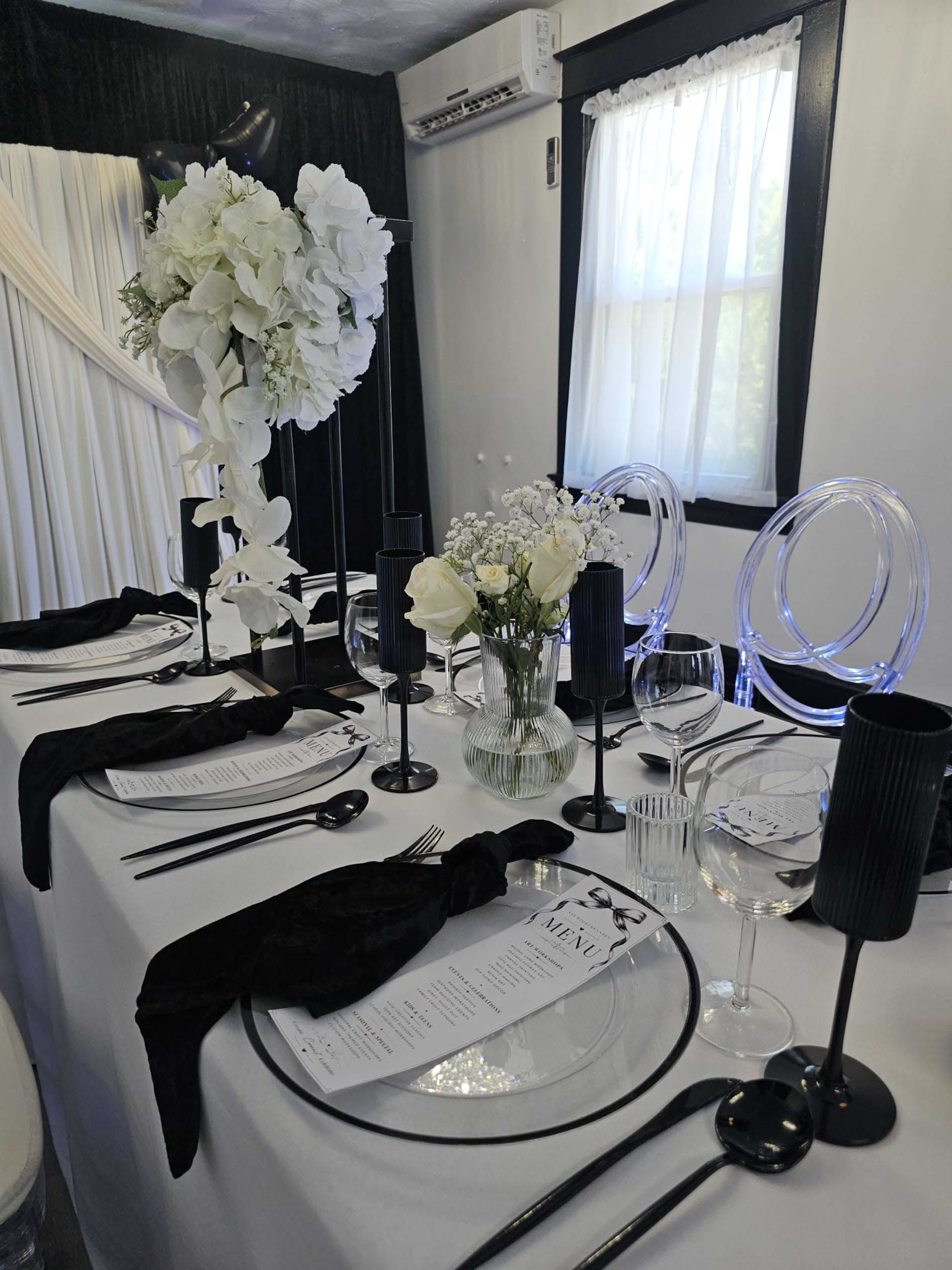 A elegantly set dining table features black tableware, clear glass accents, and floral centerpieces, surrounded by sheer curtains and black drapery.