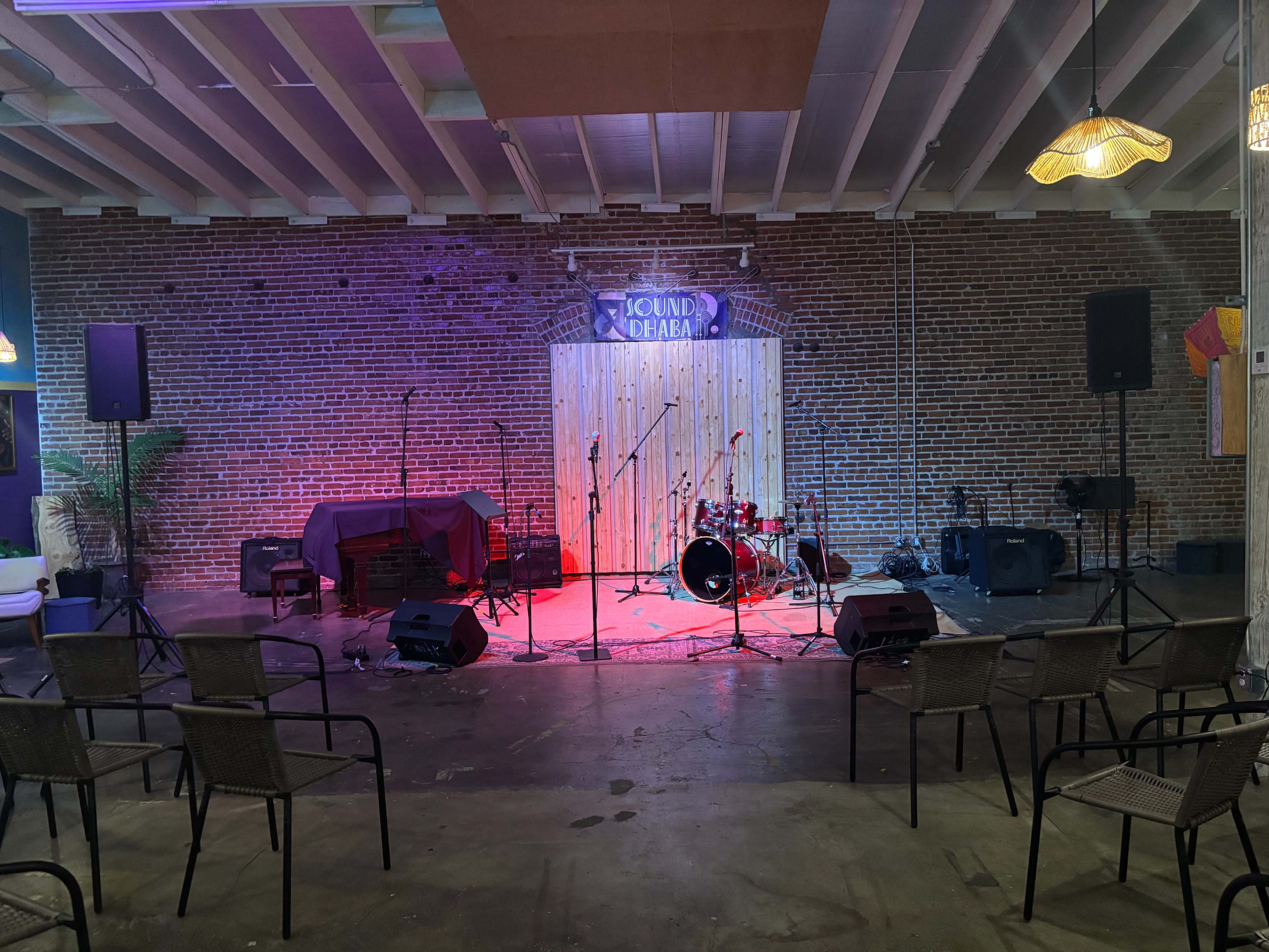 The image shows a small performance space with a stage, drum set, and sound equipment, surrounded by chairs arranged for an audience.