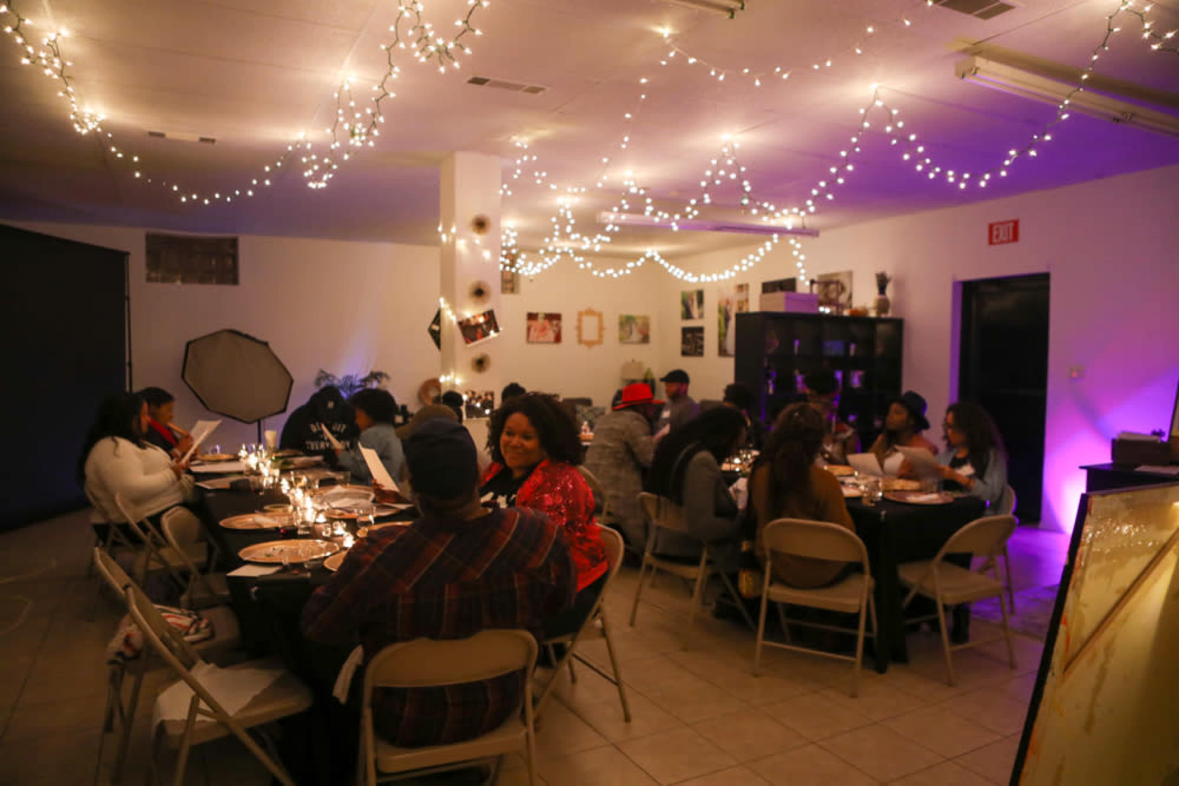A group of people is seated around tables in a dimly lit room decorated with string lights and set for a dinner gathering.