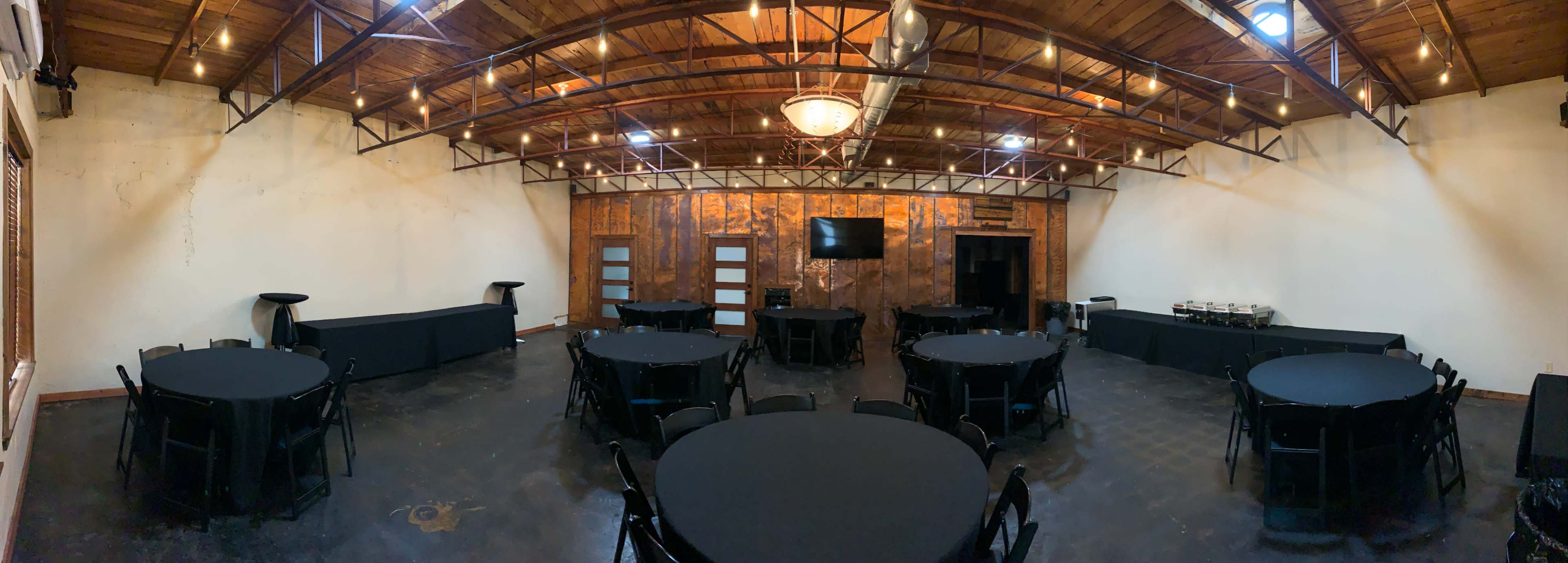 A spacious event room with black round tables arranged neatly and a wooden wall with a mounted television.