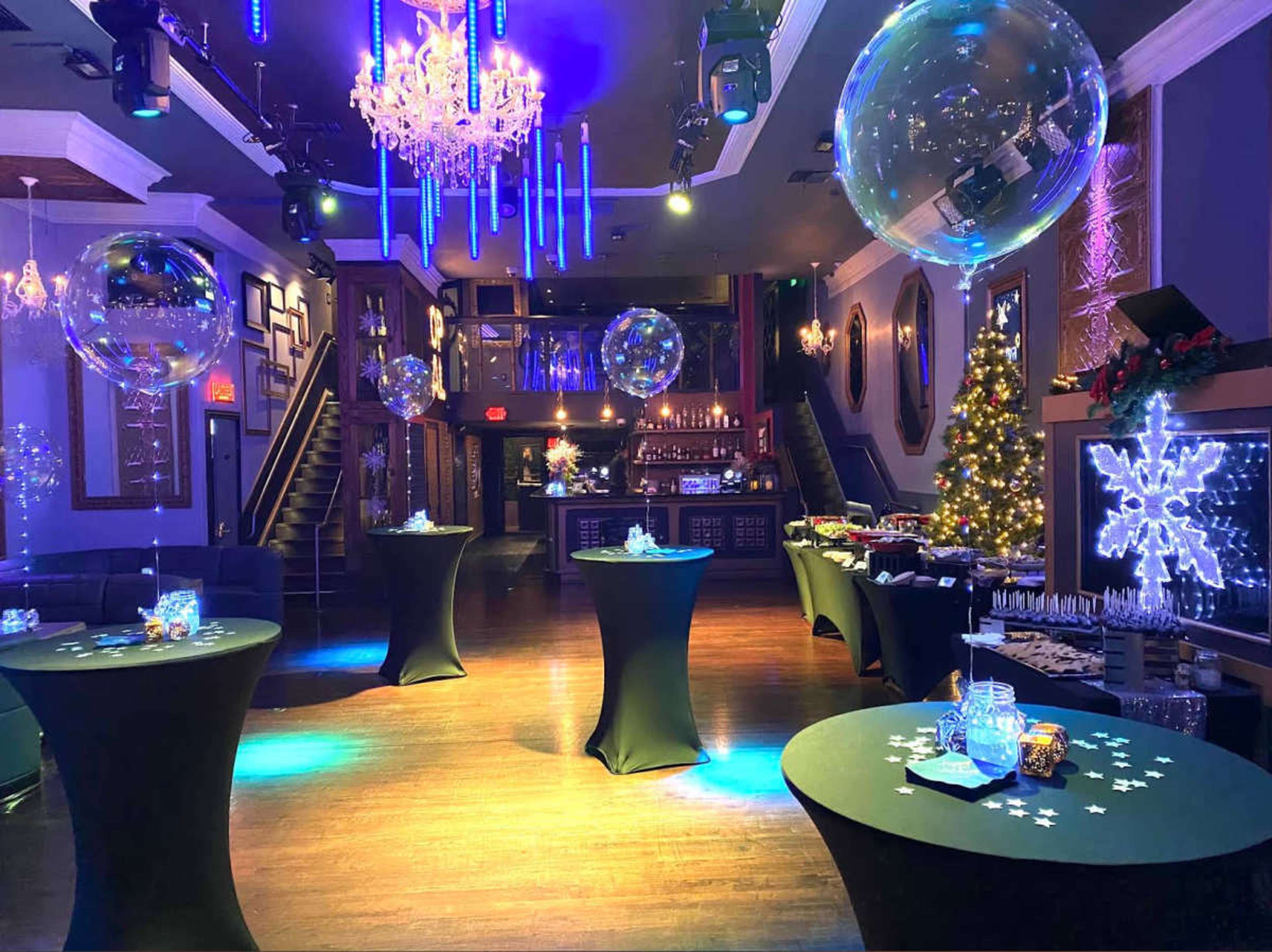 The image shows a decorated event space with tables draped in black cloth, large transparent balloons, and a Christmas tree, all illuminated by colorful lights.