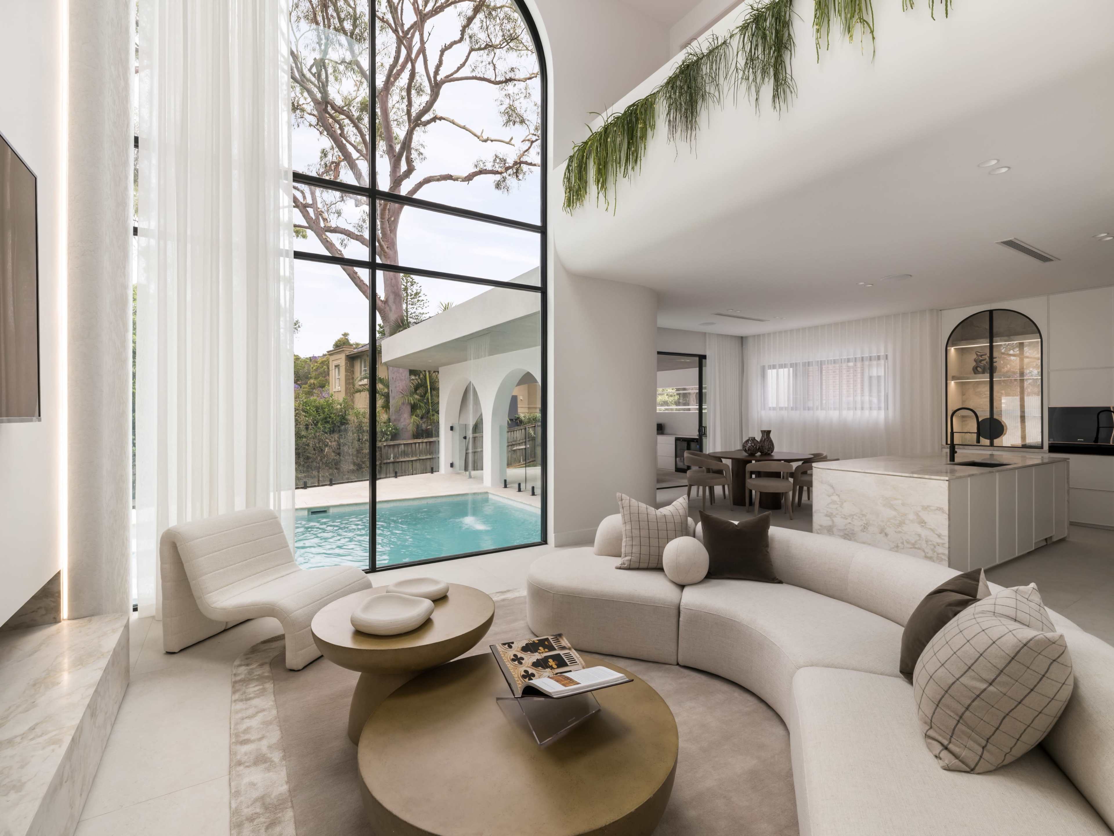 The image shows a modern, open-concept living area featuring a large curved sofa, a minimalist coffee table, and expansive windows overlooking a swimming pool surrounded by greenery.