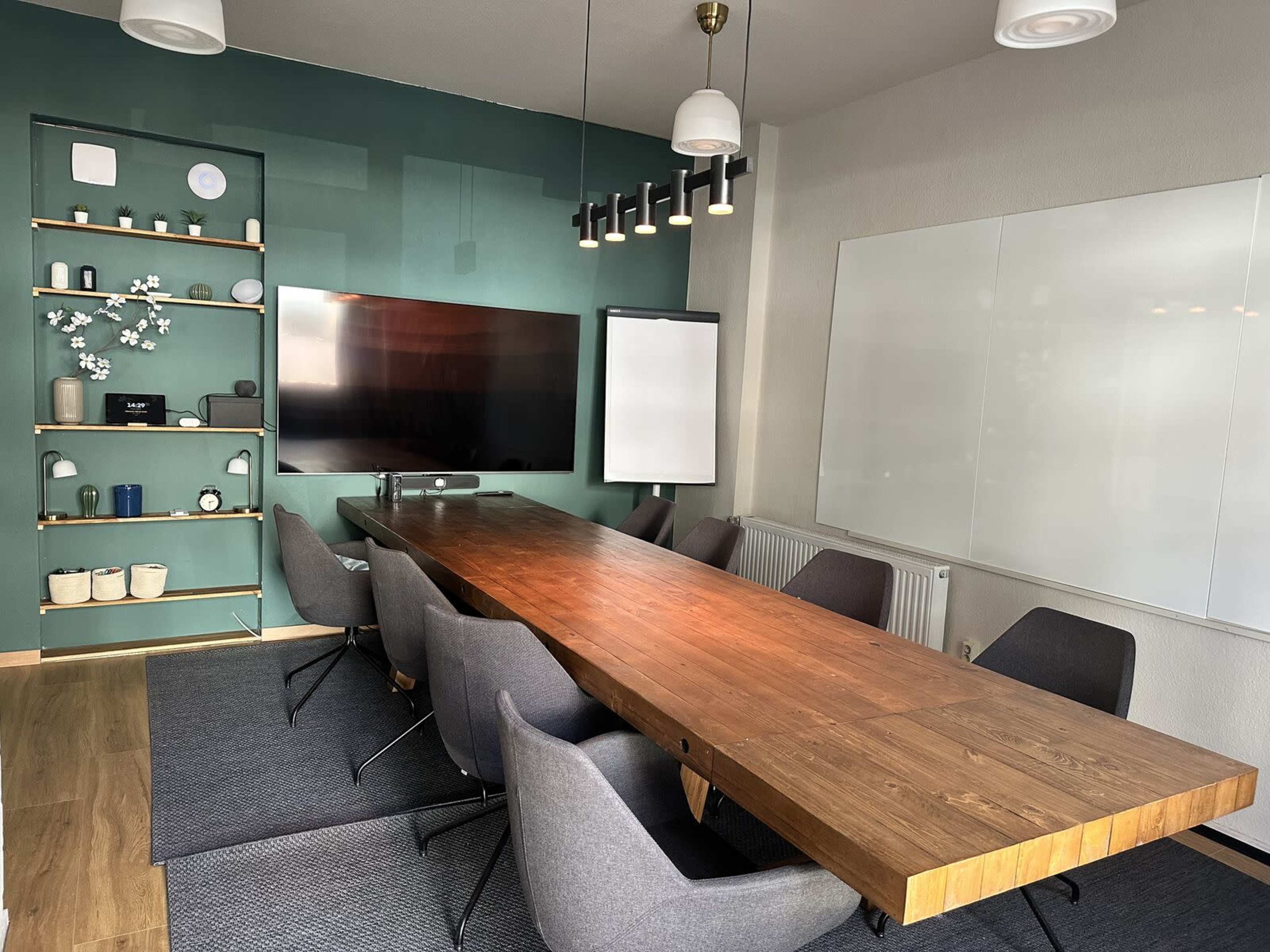 The image shows a modern conference room featuring a long wooden table, gray chairs, a large screen, and decorative shelves with various objects on a green accent wall.