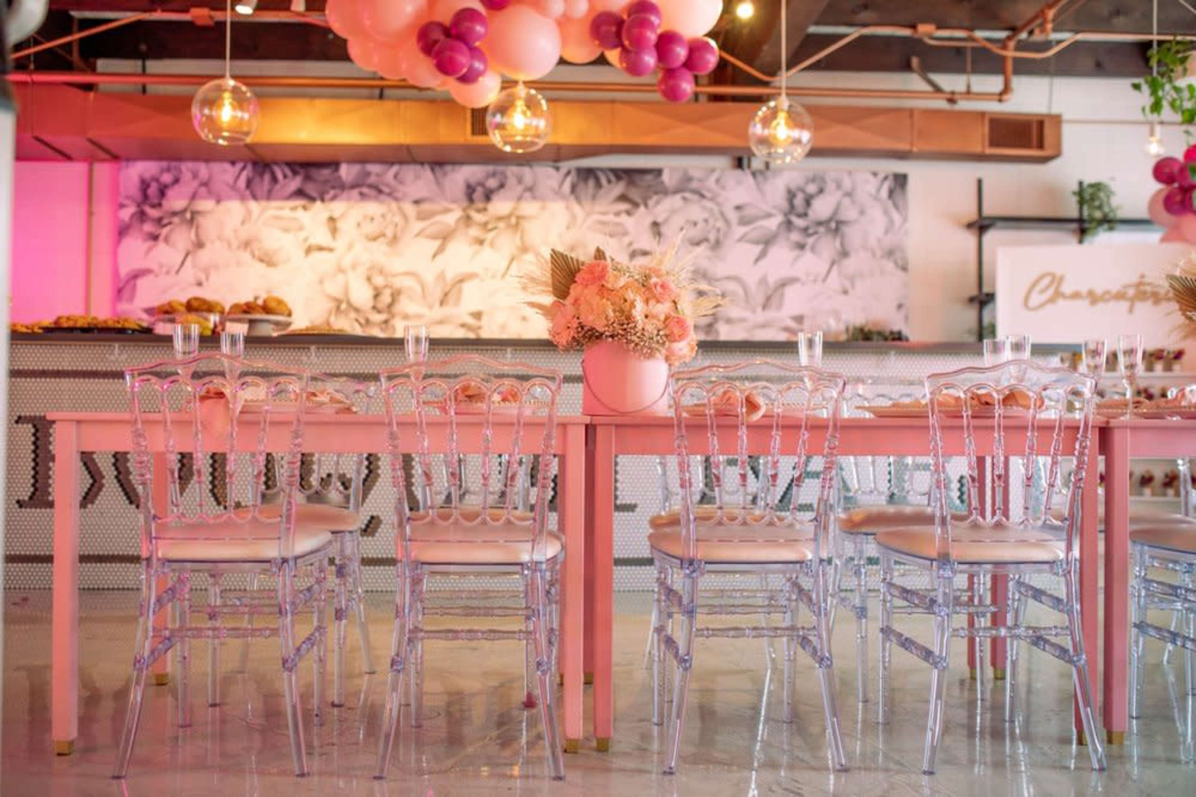 A long pink table is set with transparent chairs and adorned with a floral arrangement, while colorful balloons hang overhead in a modern event space.