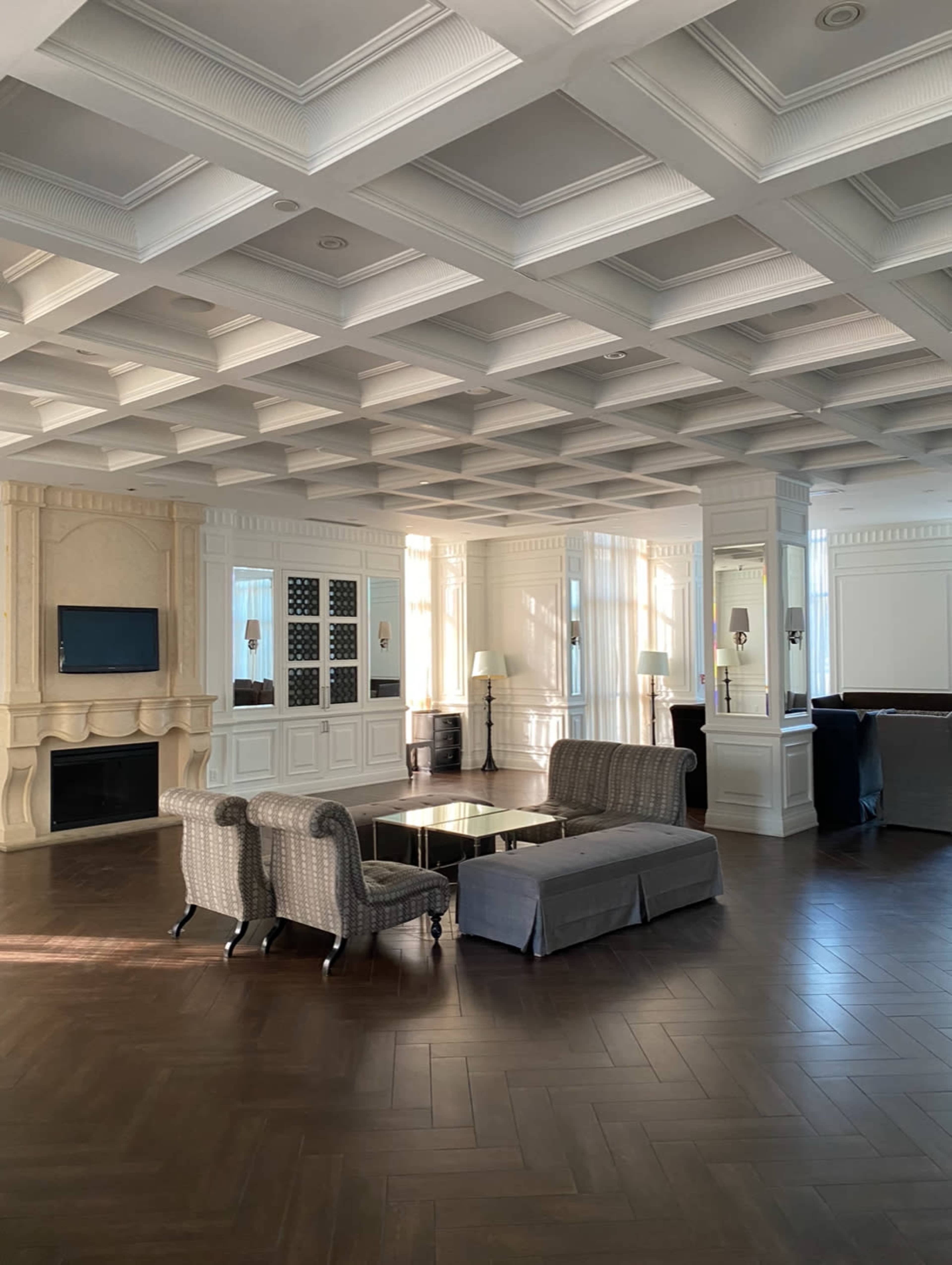 A spacious living room with coffered ceilings, elegant furniture, and natural light filtering through large windows.