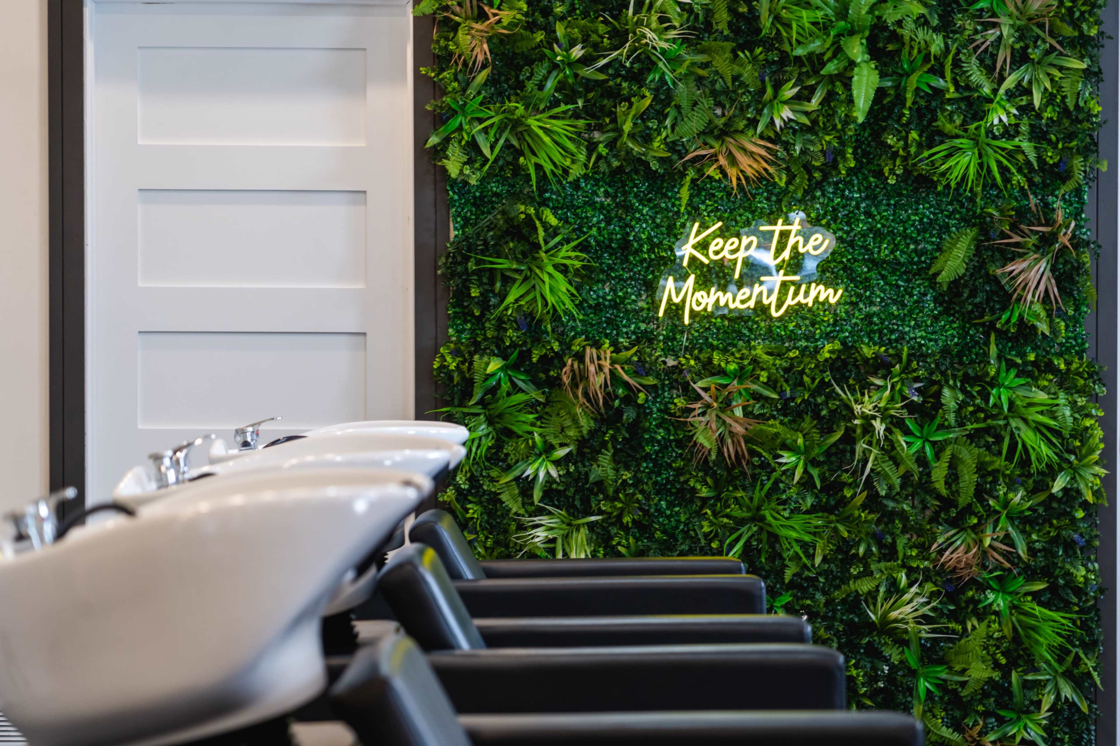 A salon interior featuring a wall covered in greenery with a neon sign that reads "Keep The Momentum," alongside several black salon chairs and wash stations.