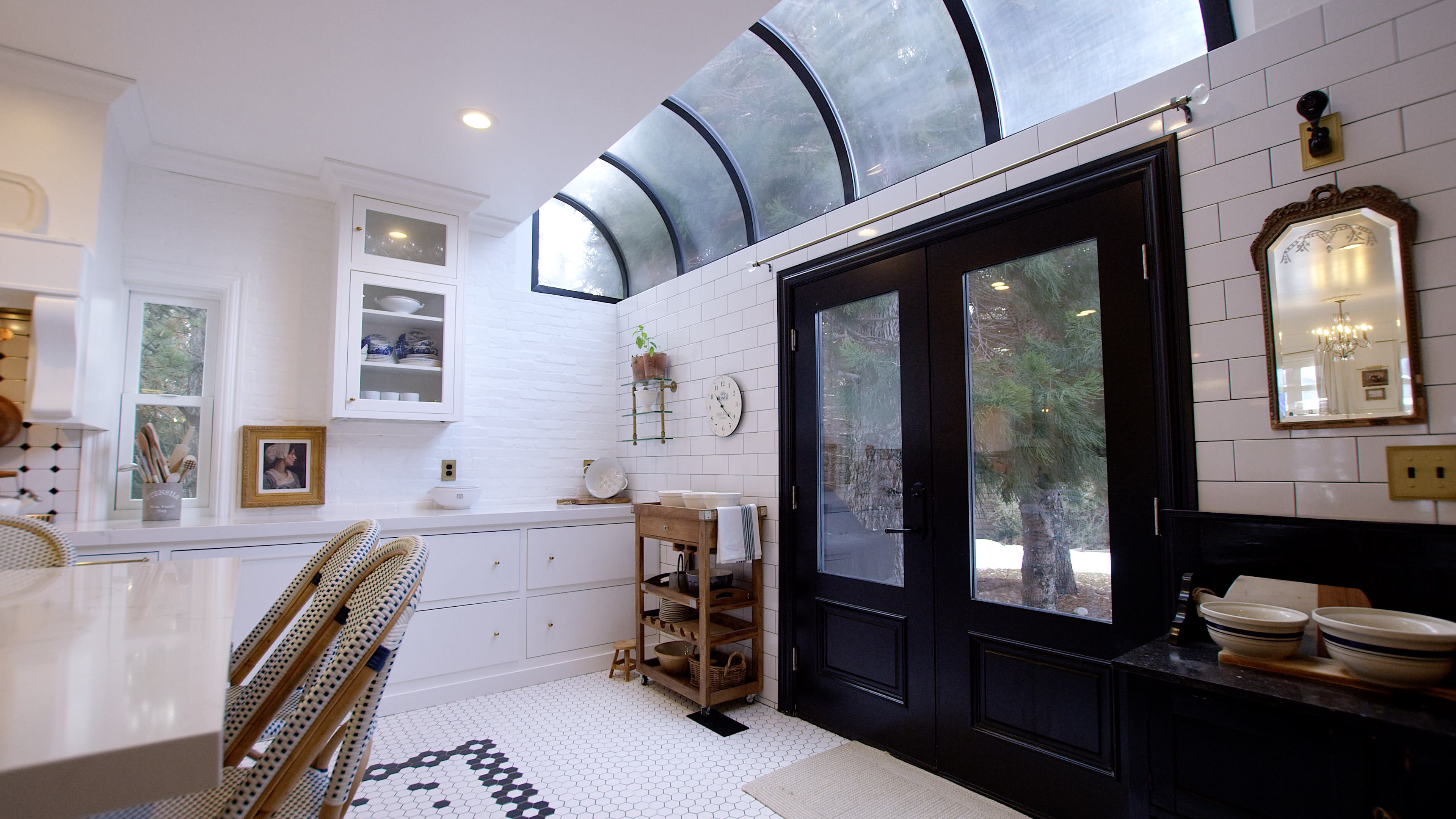A modern kitchen features white cabinetry, a hexagonal tiled floor, and large black-framed windows that overlook a garden.
