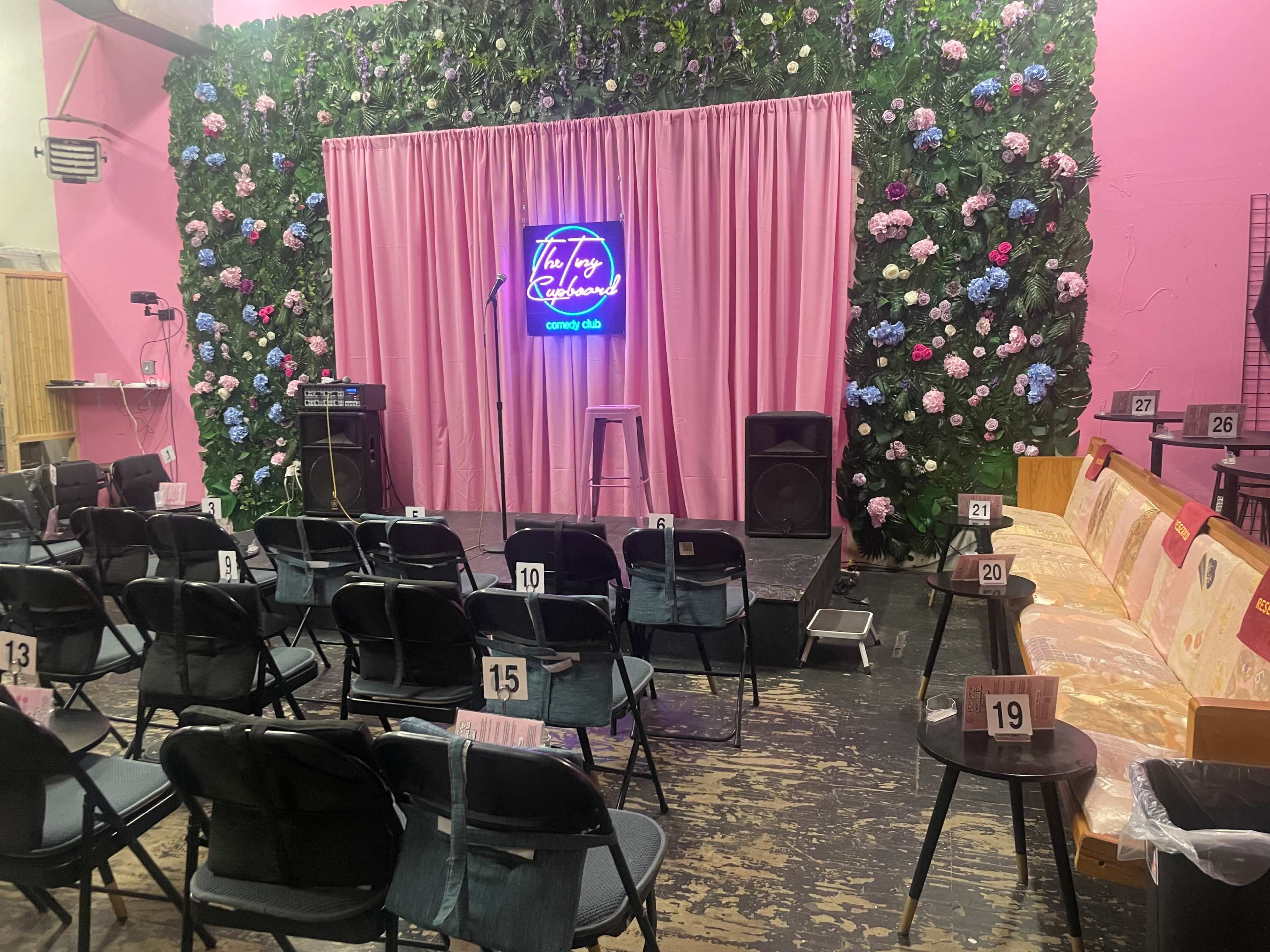 A performance stage adorned with a floral backdrop, equipped with a microphone stand, speaker equipment, and arranged seating for an audience.