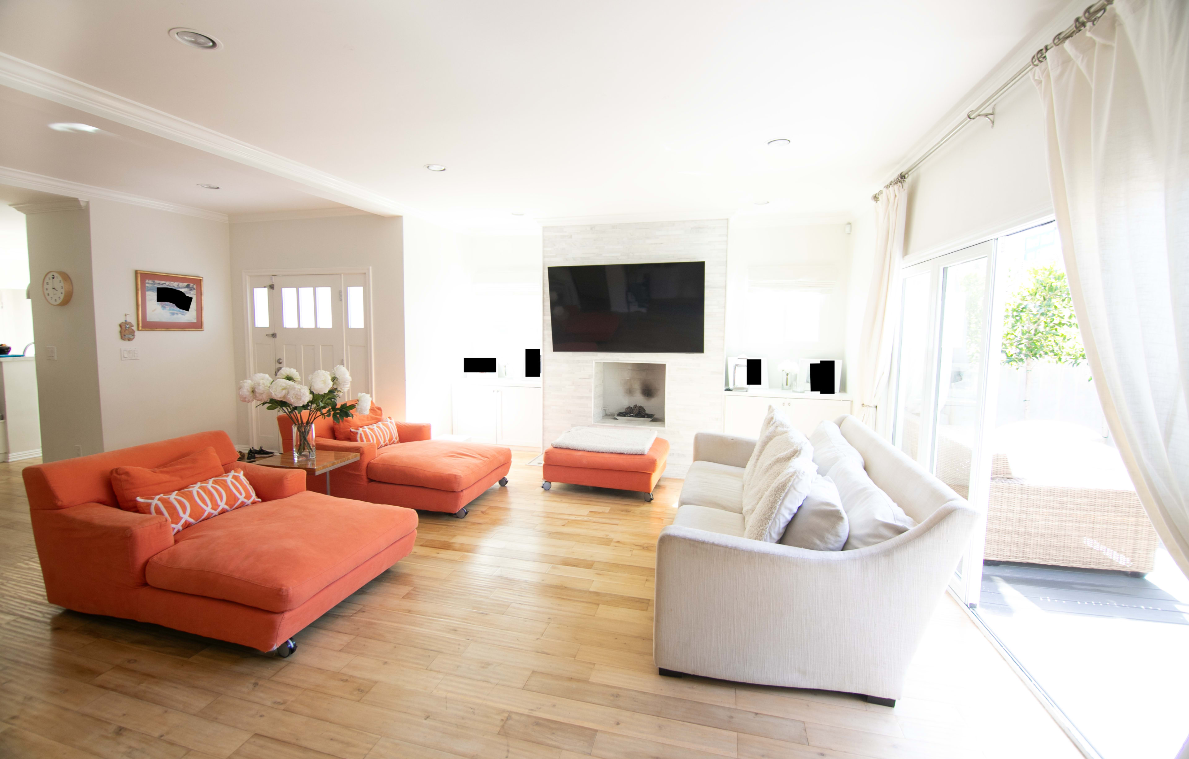 A bright living room with two orange lounge chairs and a beige sofa arranged around a white stone fireplace and a large television mounted on the wall.