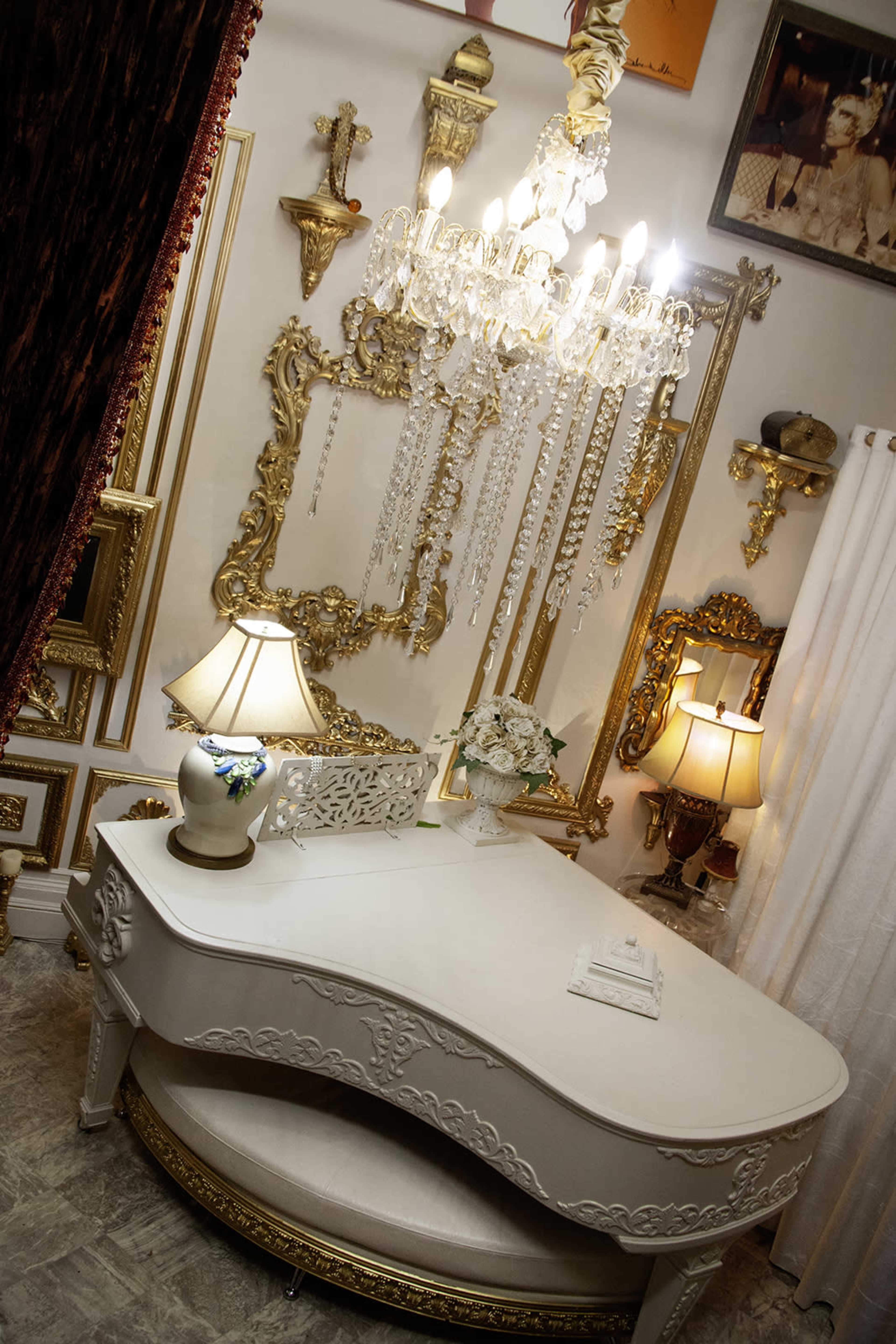 The image features an elegantly decorated room with a white, curved desk, two lamps, and a chandelier surrounded by ornate gold-framed wall decorations.