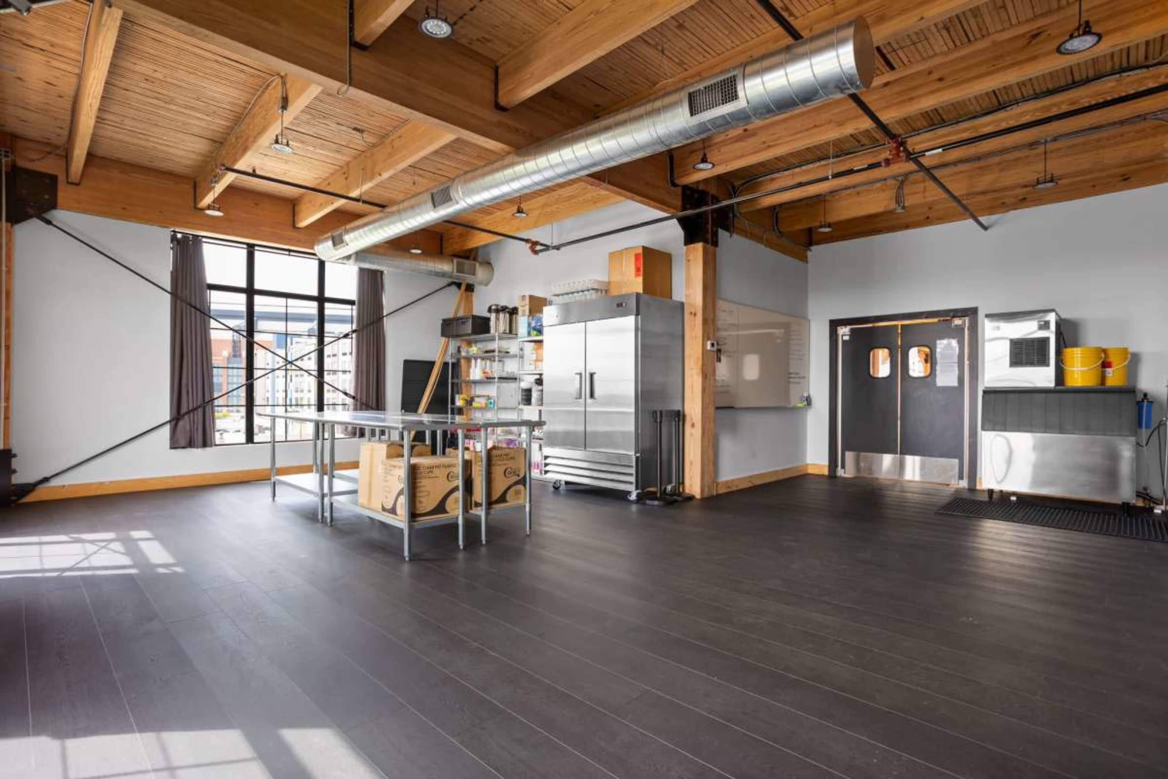 The image shows an industrial-style interior space with exposed wooden beams, a large window, a stainless steel kitchen setup, and a polished black floor.