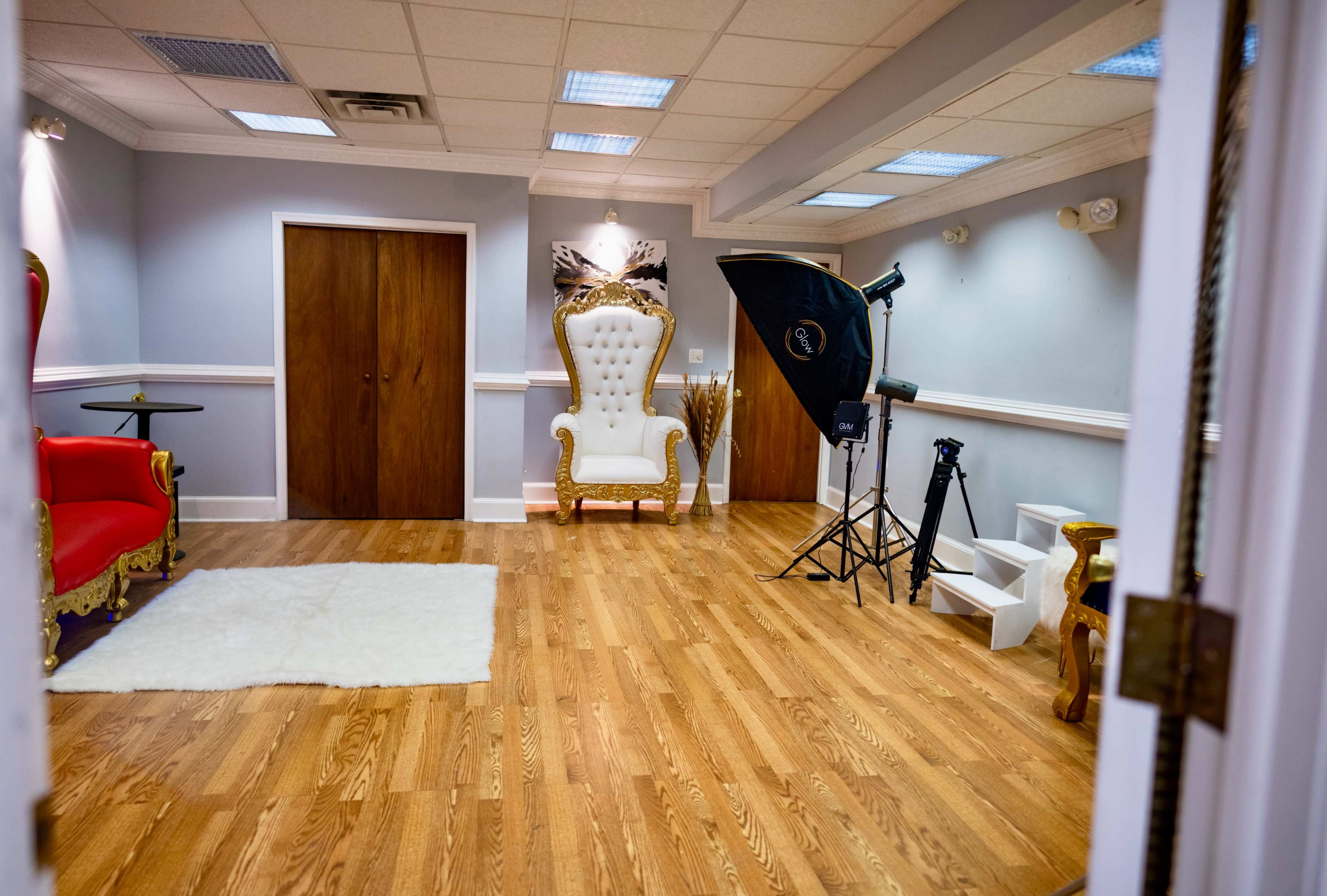 A spacious studio with wooden flooring, featuring a large royal-style white chair, a red chair, and photography equipment set up in the corner.