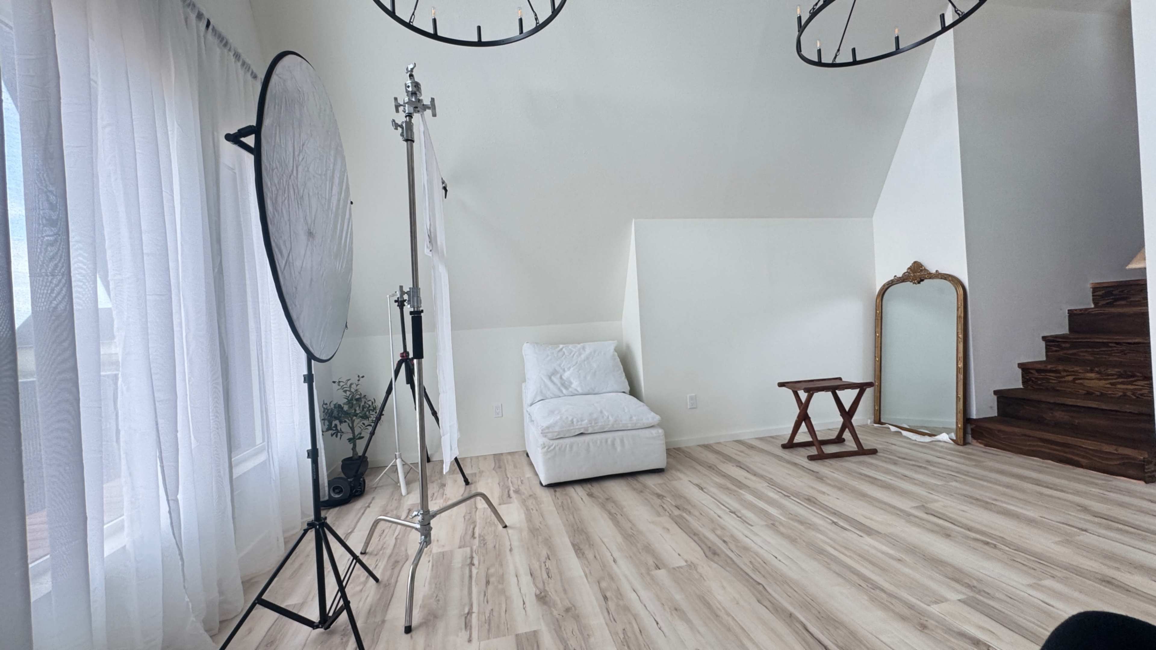 The image shows a bright, minimalist indoor space featuring a white chaise lounge, a mirror, a small wooden table, and a stand with a light reflector near large windows.
