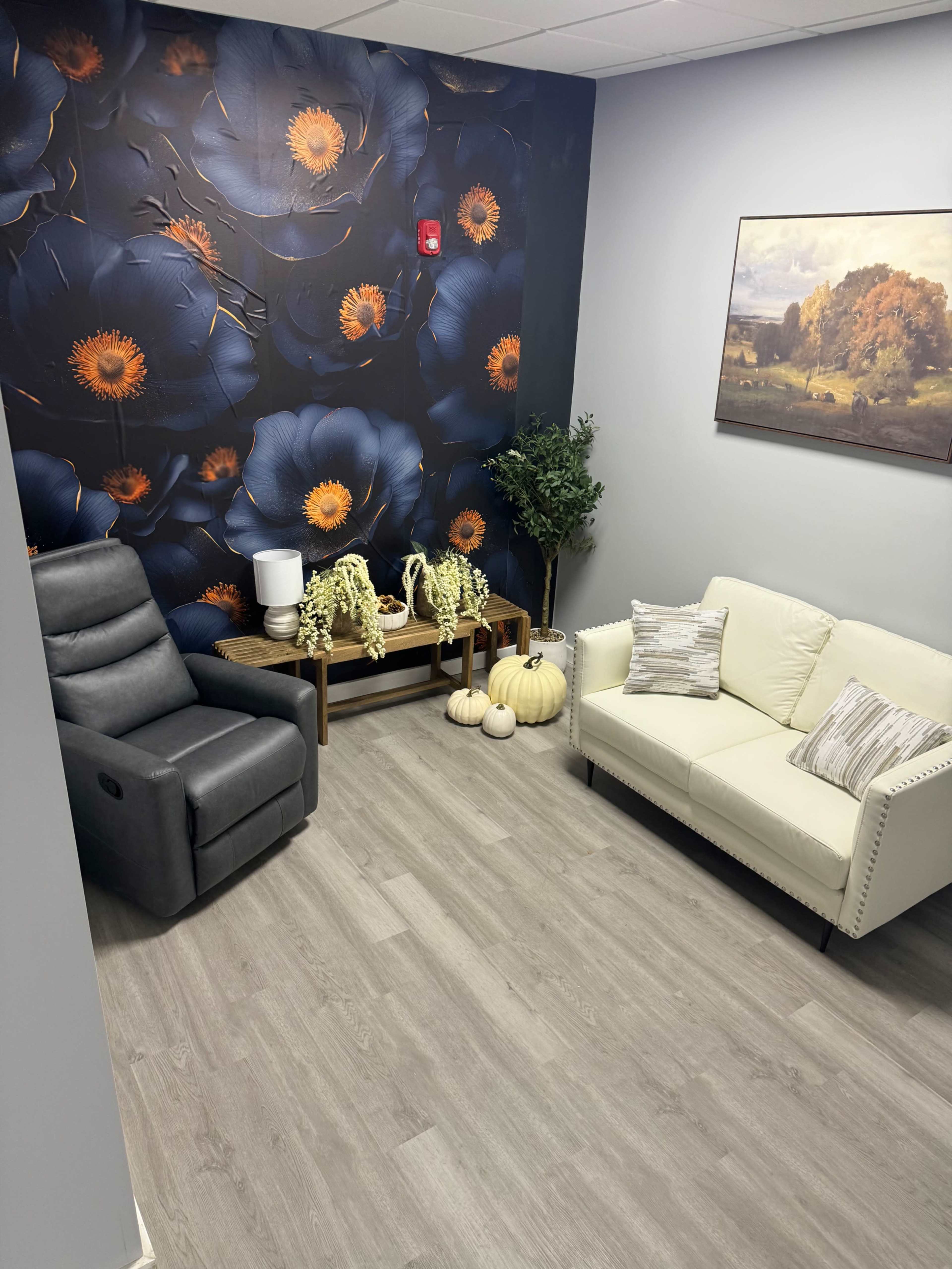 The image shows a small sitting area with a gray recliner, a white sofa, a wooden table with decorative items, and a wall adorned with large floral wallpaper.