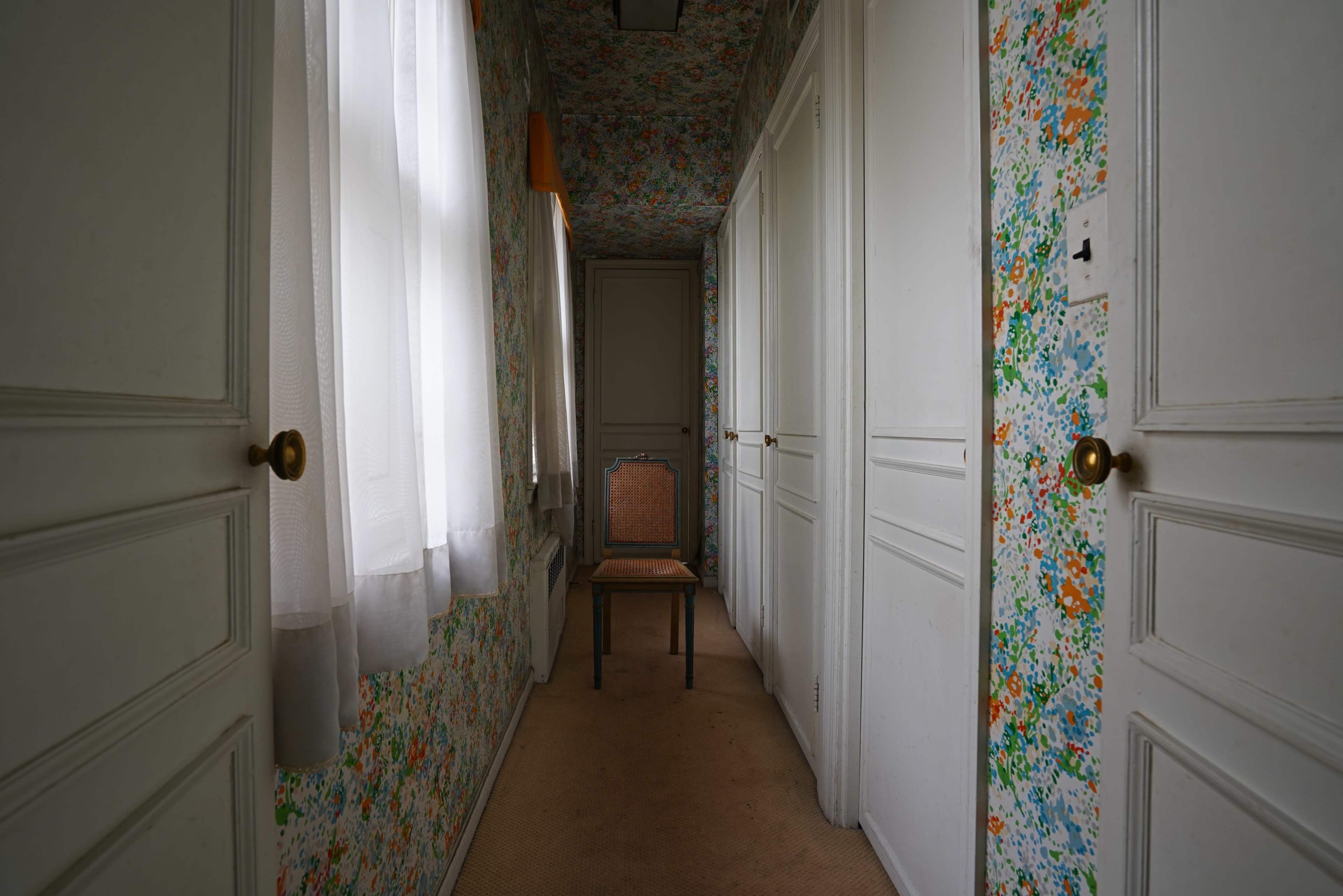 A narrow hallway is lined with floral wallpaper and features a single wooden chair in front of a closed door.