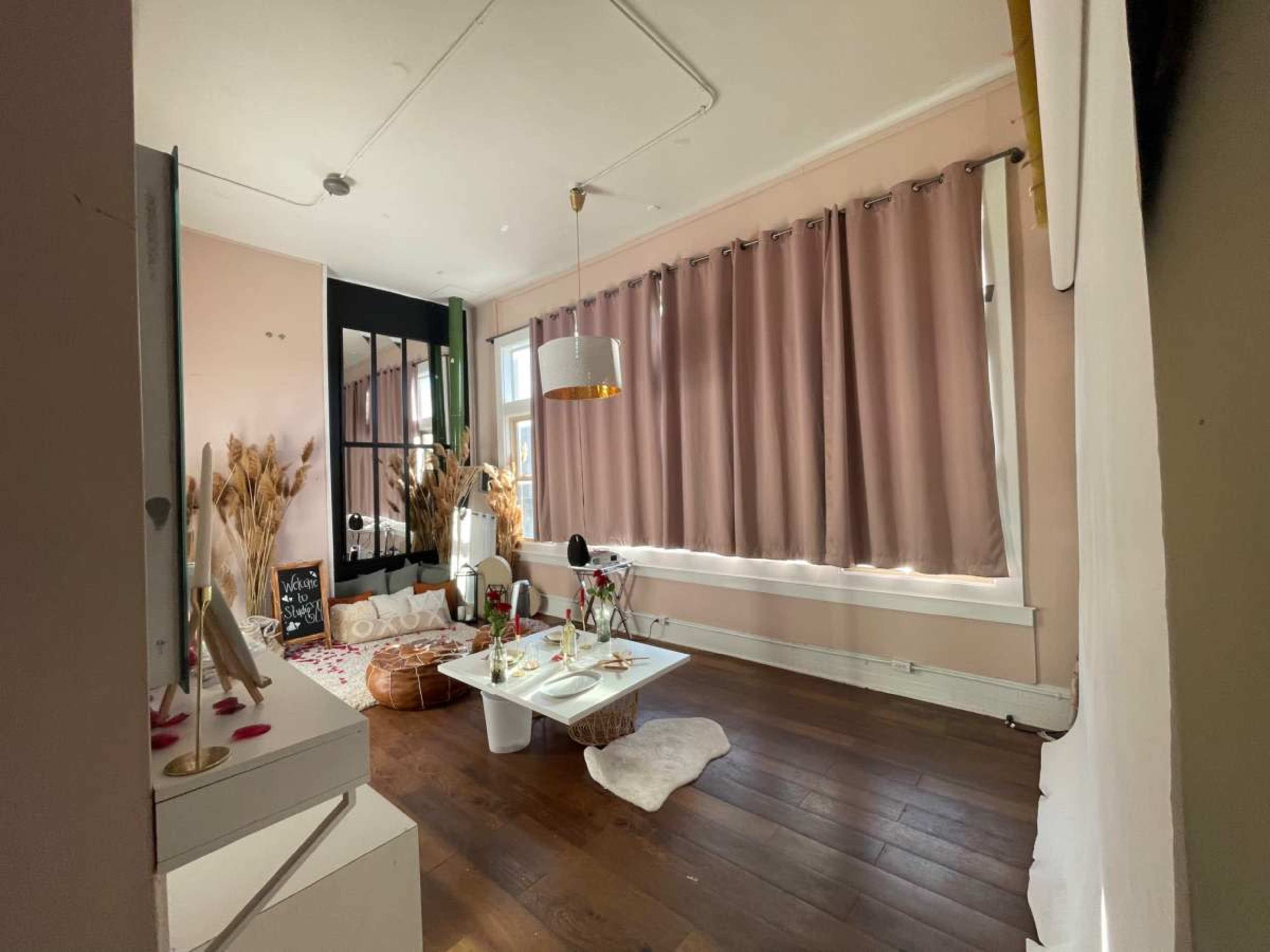 A cozy interior space features a large mirror, soft curtains, a low white table set with decor, and wooden flooring.