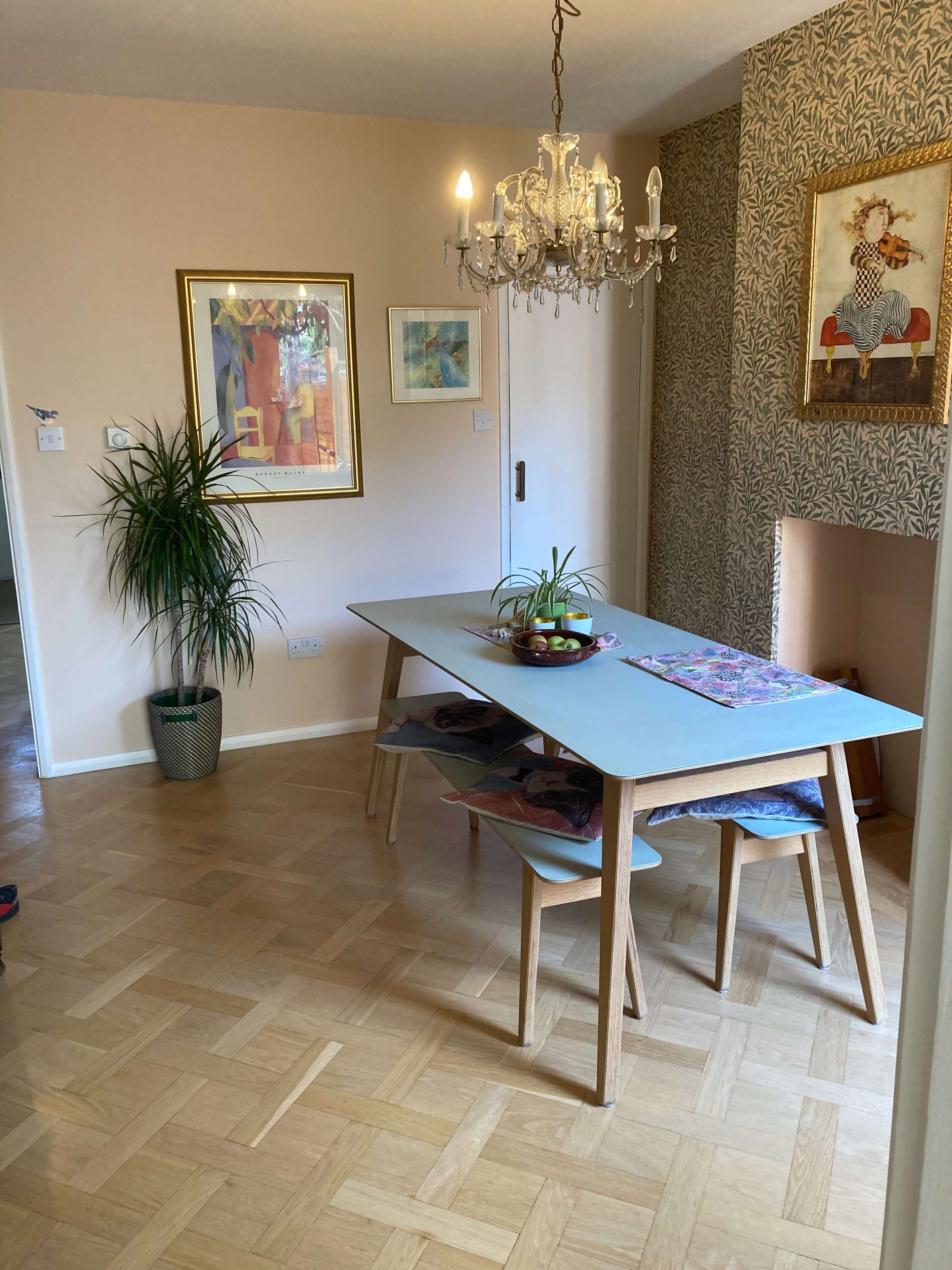 A light-filled dining area featuring a wooden table surrounded by chairs, a chandelier overhead, and wall decor including framed artwork and patterned wallpaper.