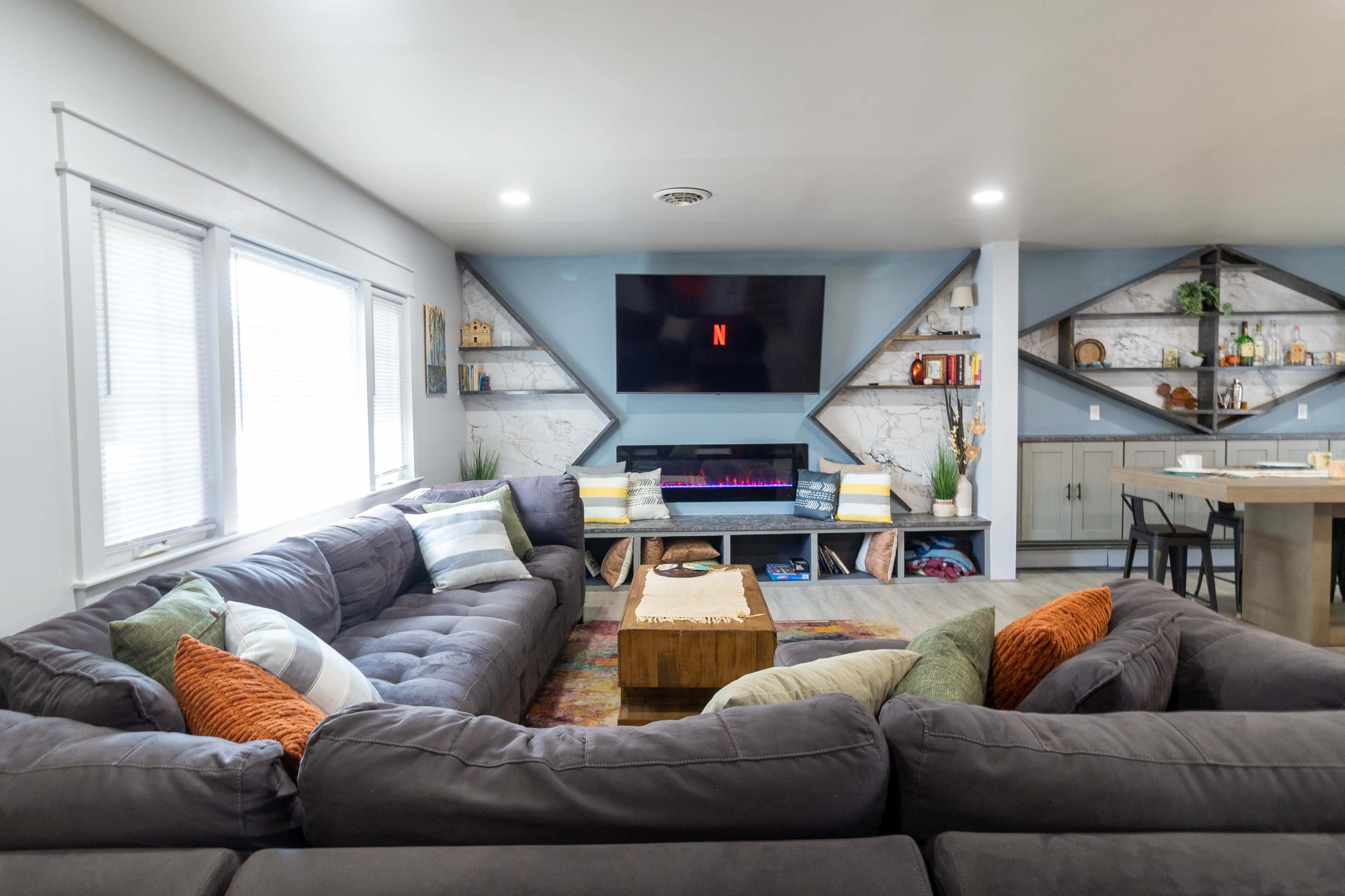A modern living room with a large sectional sofa, a mounted TV displaying the Netflix logo, and shelves filled with decorative items and drinks.