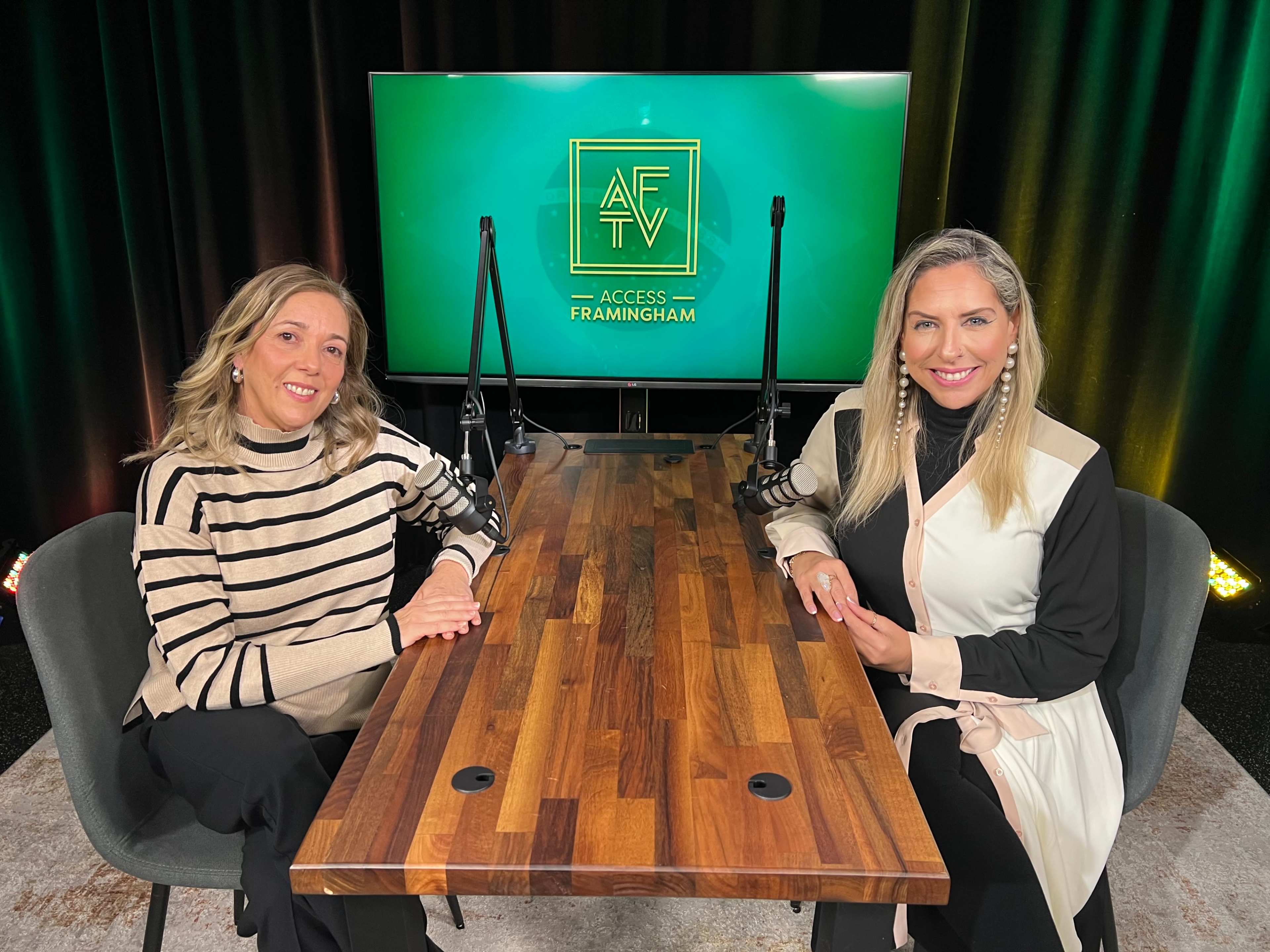 Two women sit at a wooden table in front of a green backdrop displaying the logo for Access Framingham TV.