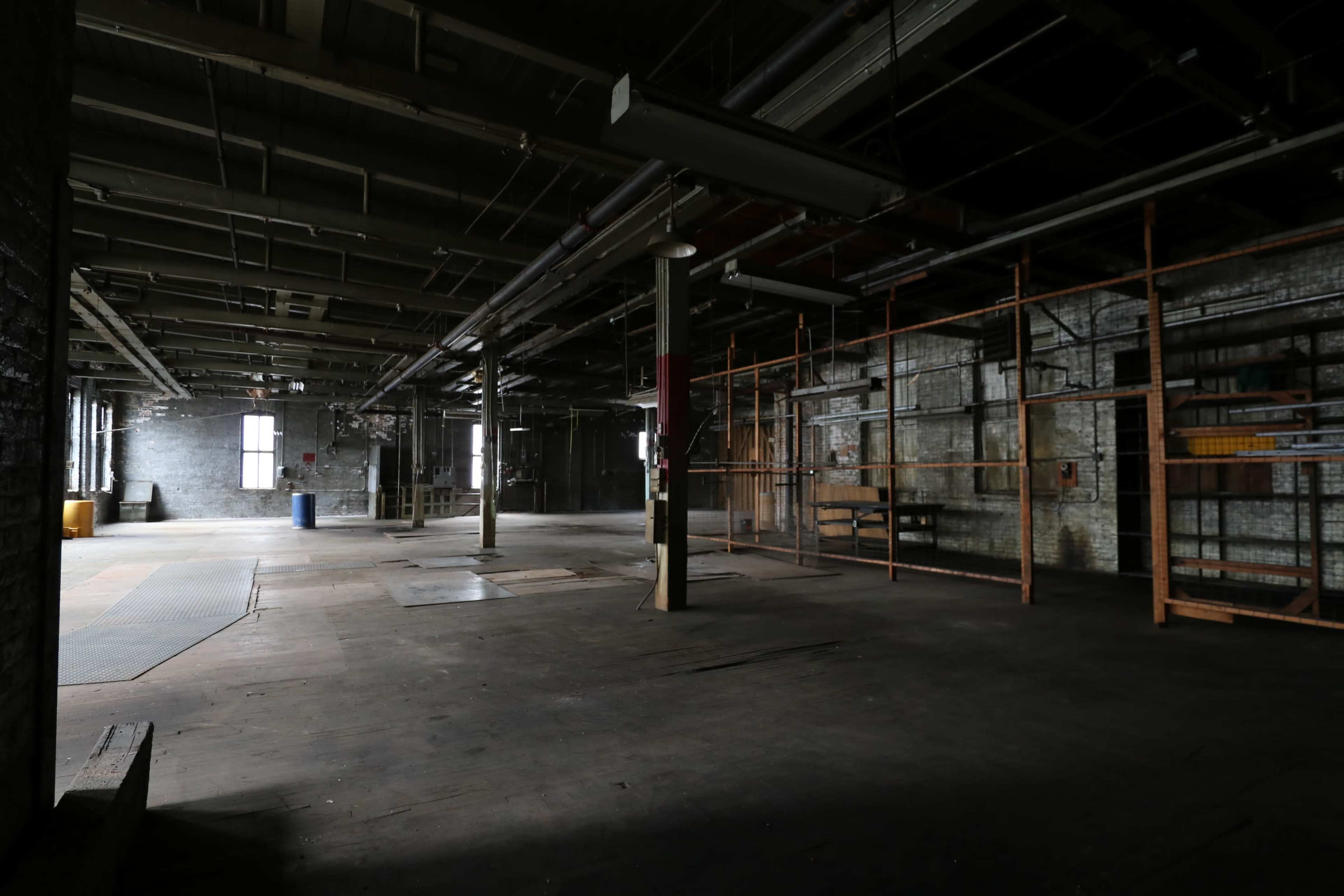 The image shows an empty industrial warehouse with exposed beams, large windows, and scattered construction materials.